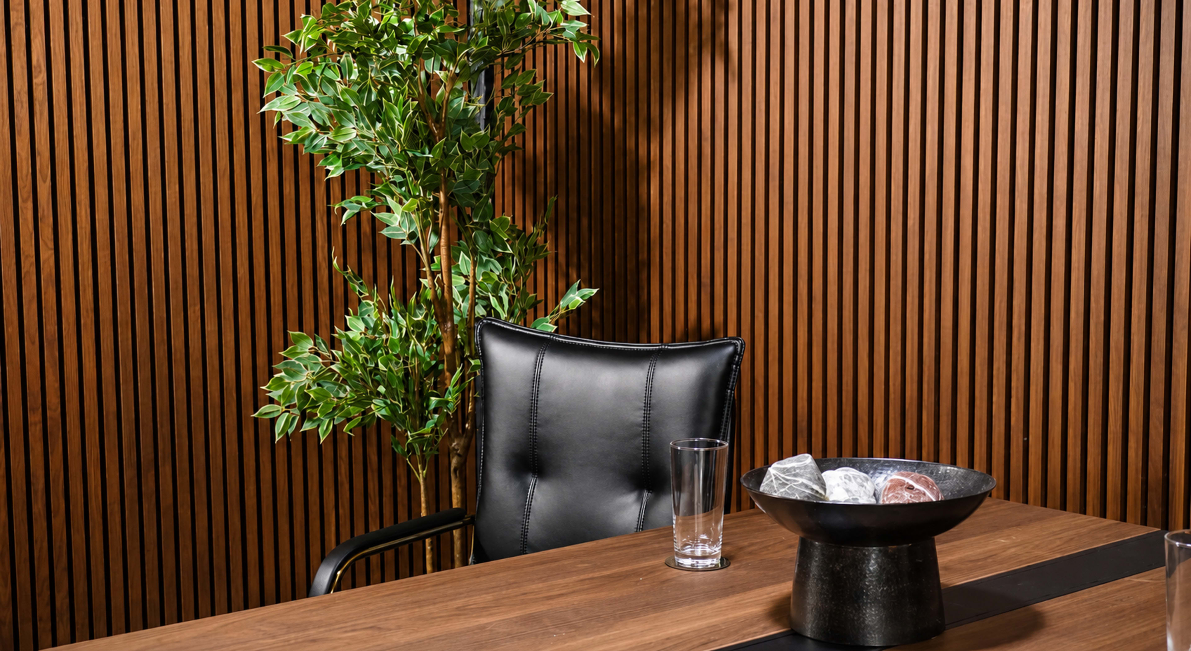 A black leather chair is positioned next to a wooden table featuring a bowl with decorative stones, alongside a clear glass. A leafy green plant is placed against a backdrop of vertical wooden slats.