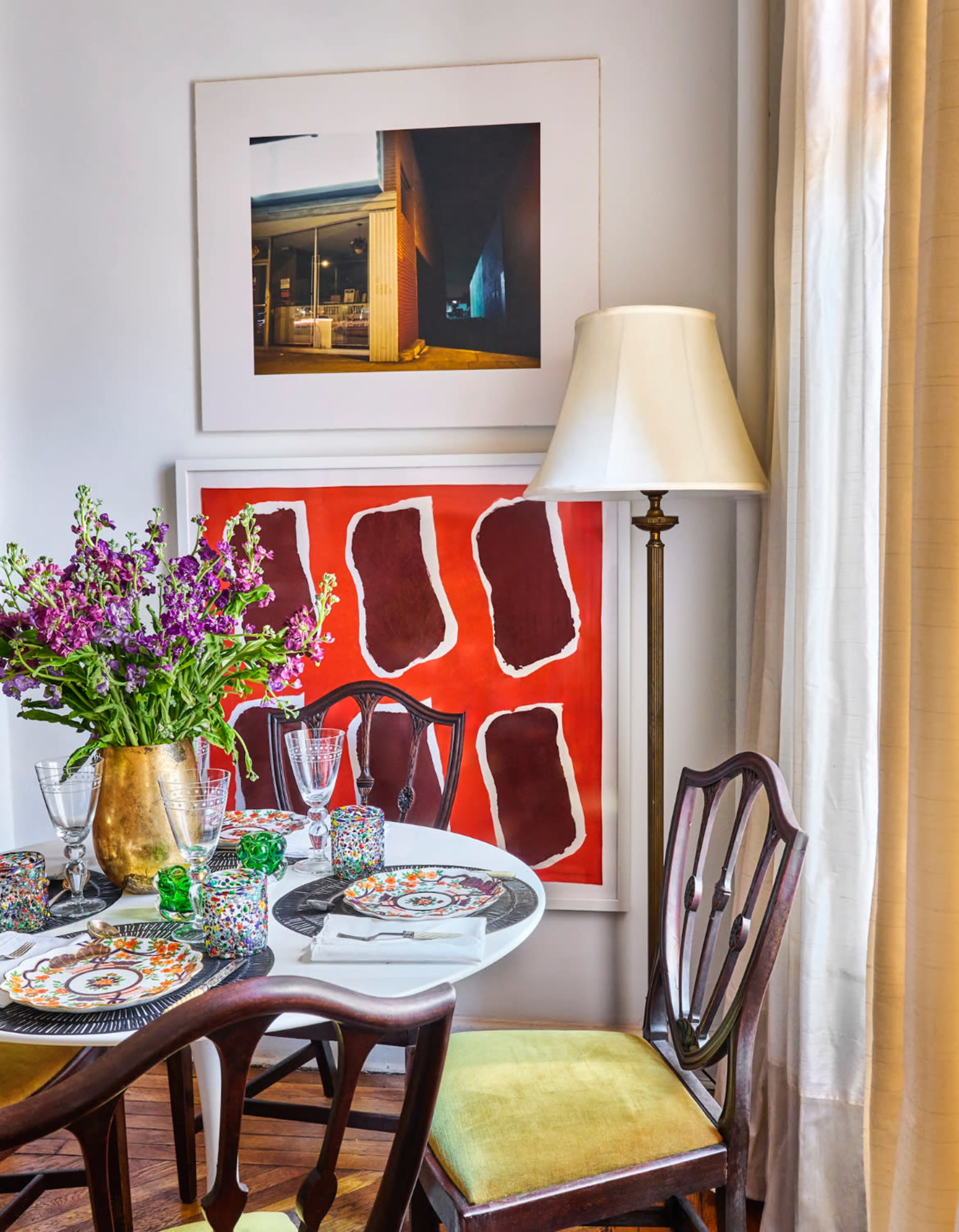 A dining table set with decorative plates and glasses sits next to a vibrant piece of wall art and a floor lamp, surrounded by flowers in a vase.