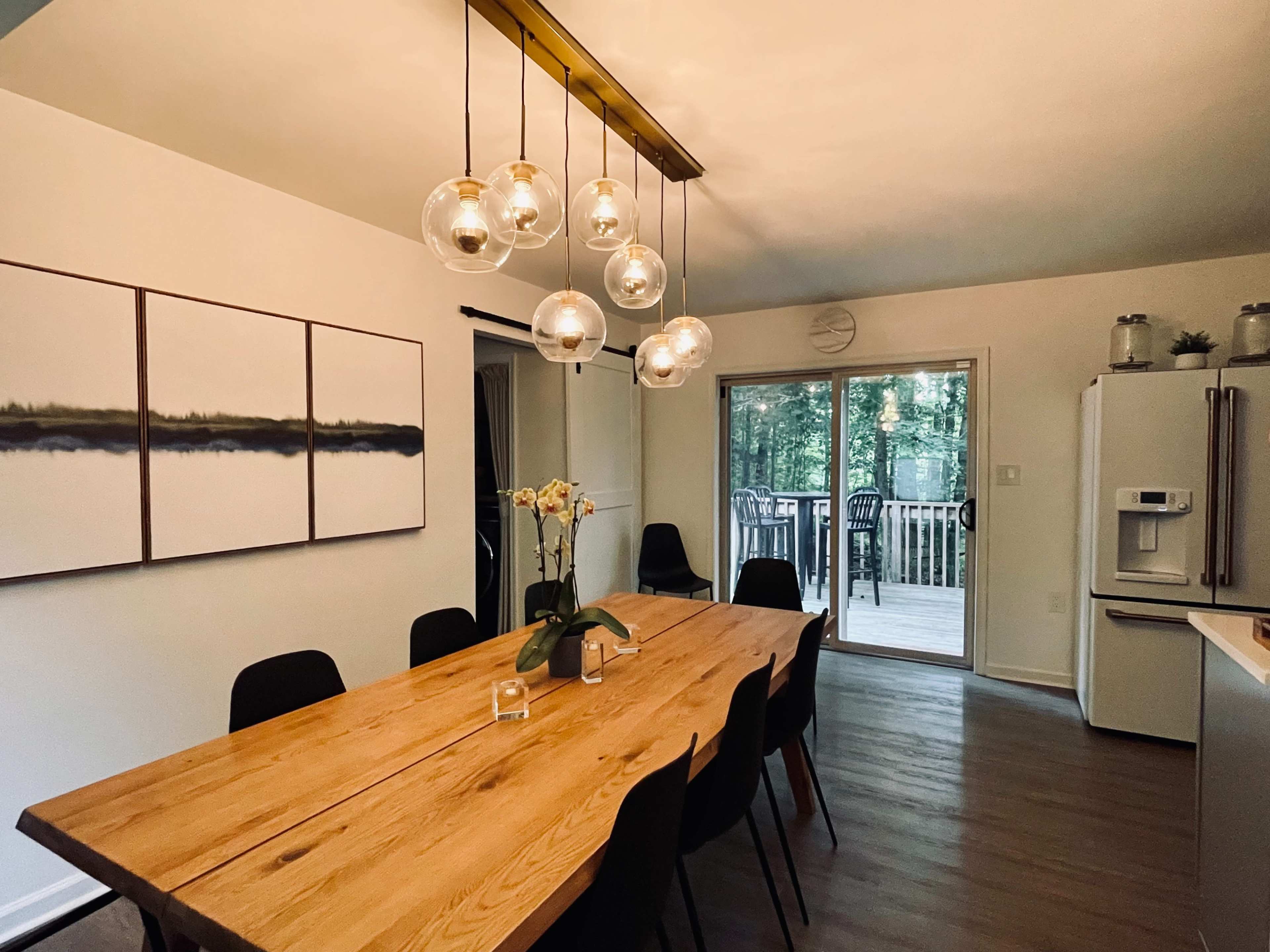 A dining area features a wooden table surrounded by black chairs, with pendant lights overhead and sliding doors leading to a deck in the background.