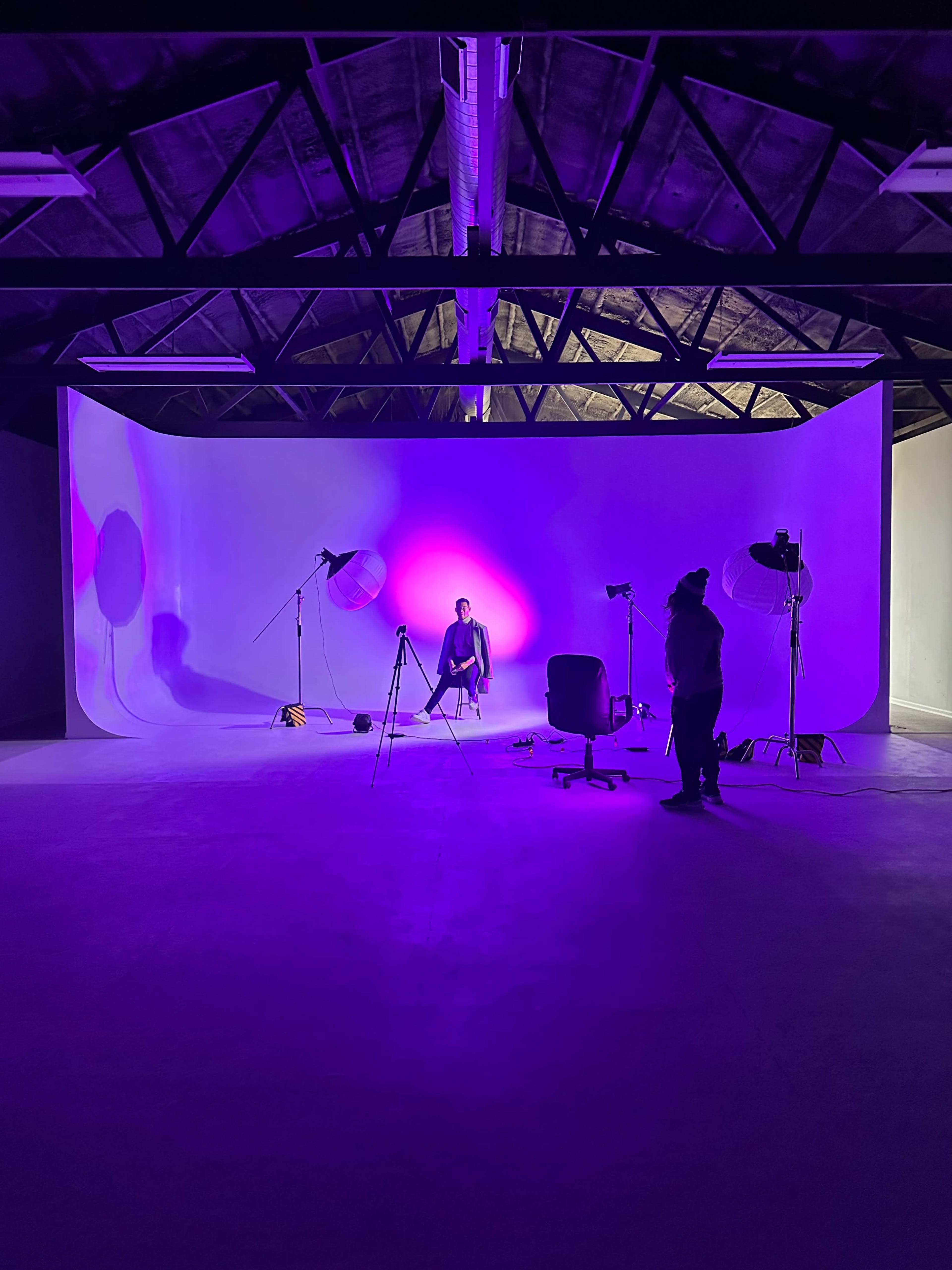 A well-lit studio space features a model sitting against a backdrop with purple lighting, surrounded by photography equipment.