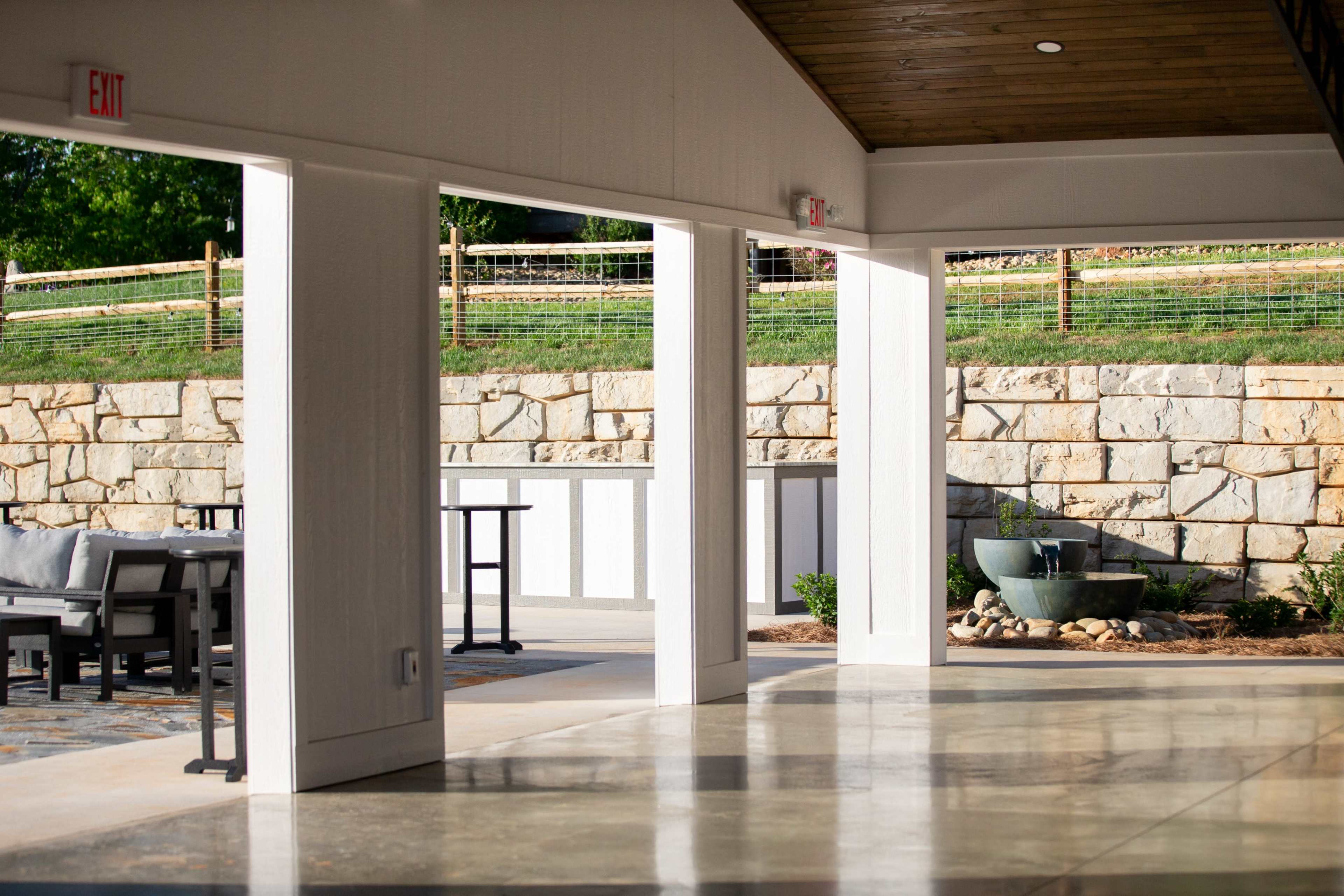 A spacious, modern indoor area with large openings leading to an outdoor space featuring a stone wall and a decorative fountain.