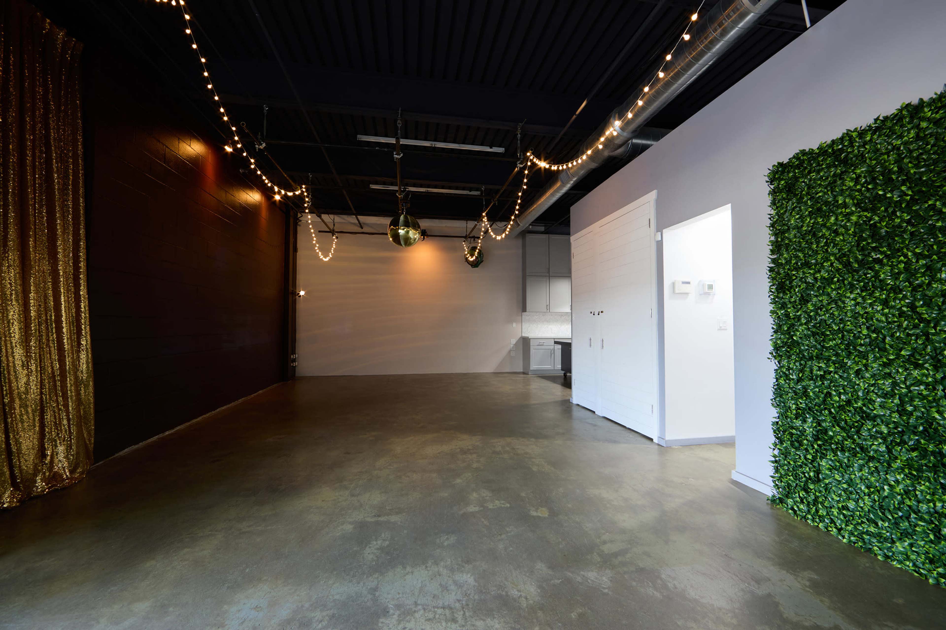 The image shows a spacious industrial room with a polished concrete floor, one wall draped in gold fabric, and another adorned with a green wall of artificial foliage.