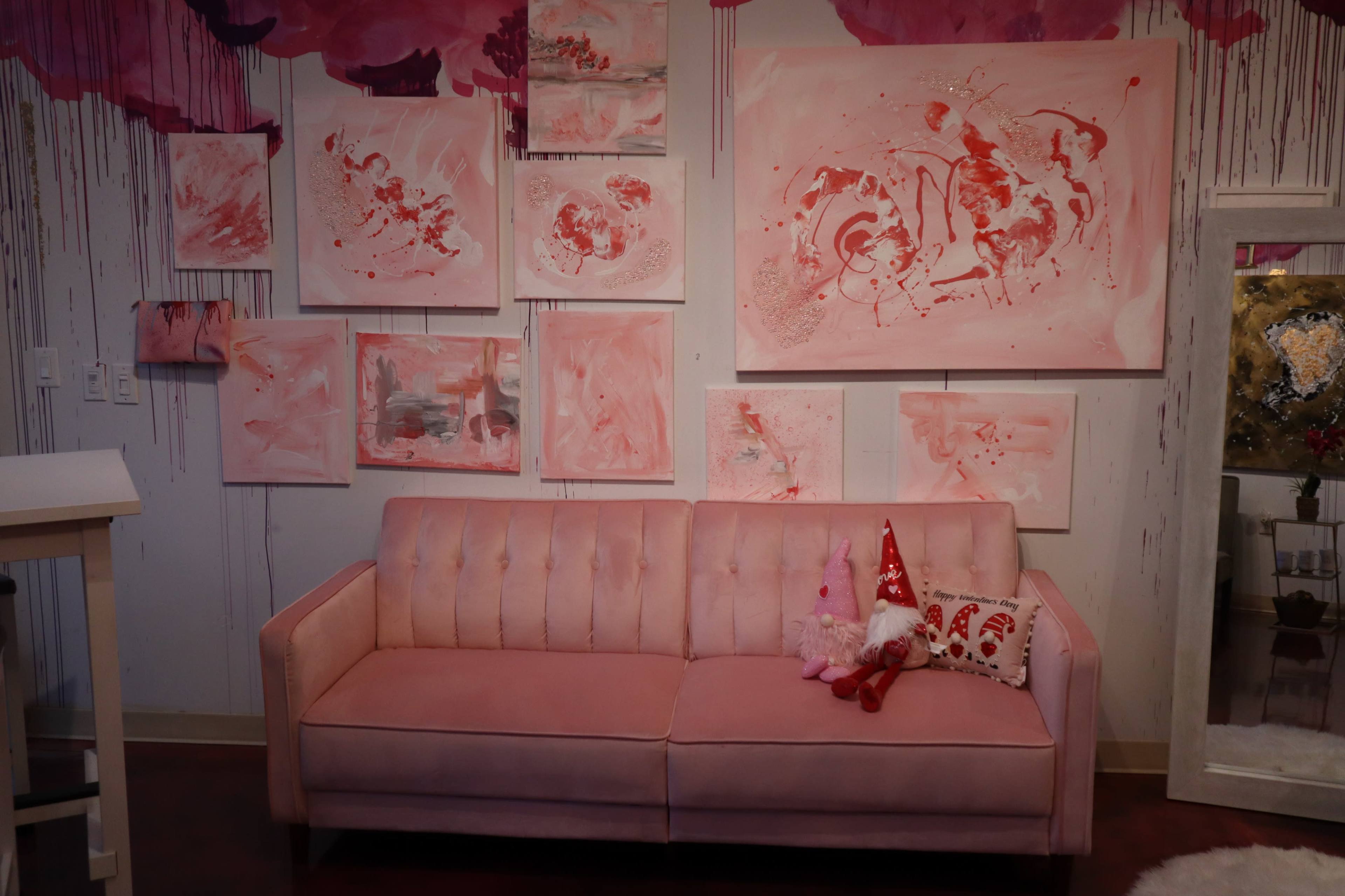 A pink couch is positioned against a wall adorned with various pink abstract paintings, complemented by a decorative pillow and plush toys.
