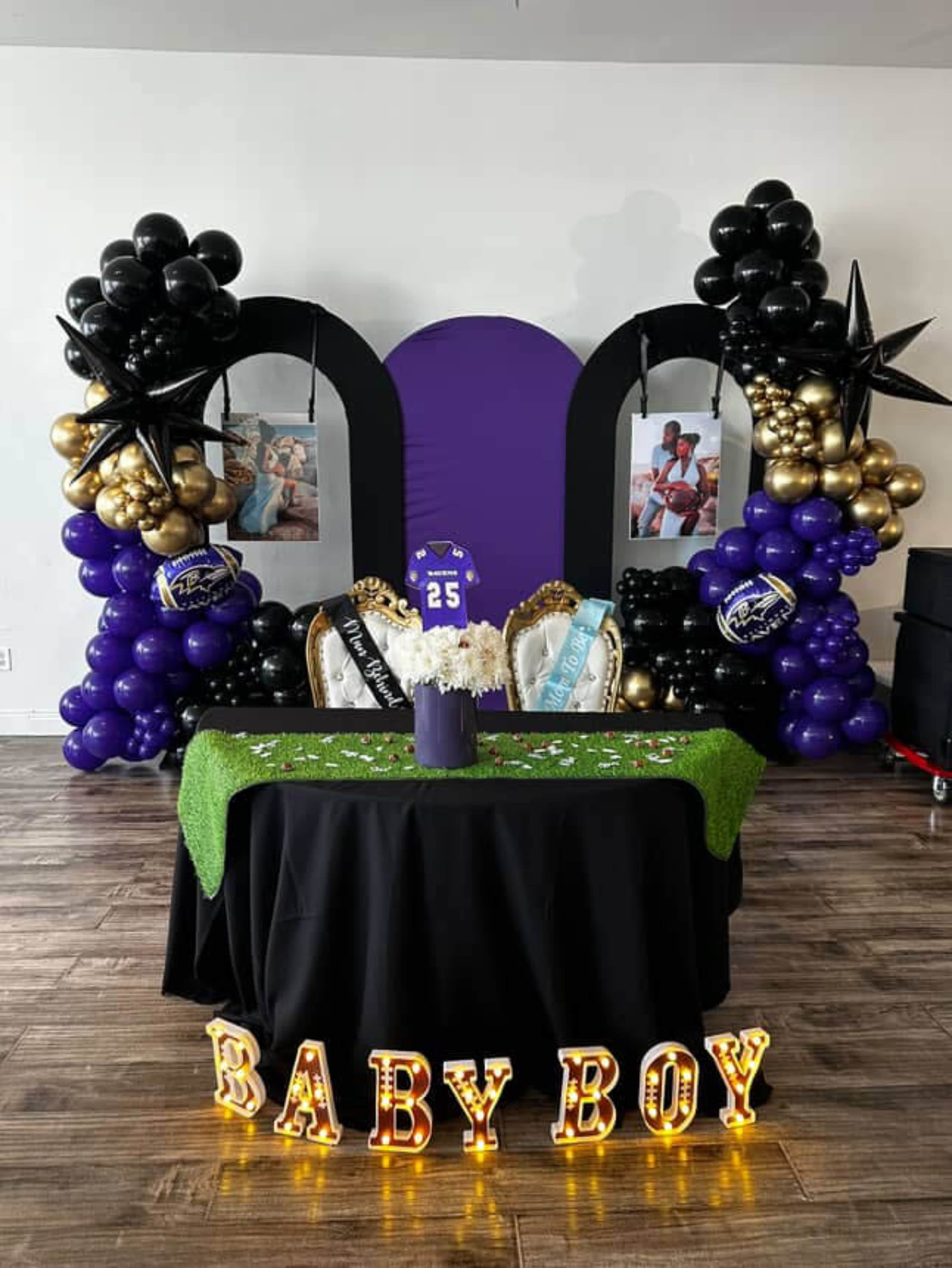The image shows a decorated event space featuring a table with a green tablecloth, white flowers, and a "BABY BOY" light-up sign, surrounded by black, gold, and purple balloons.