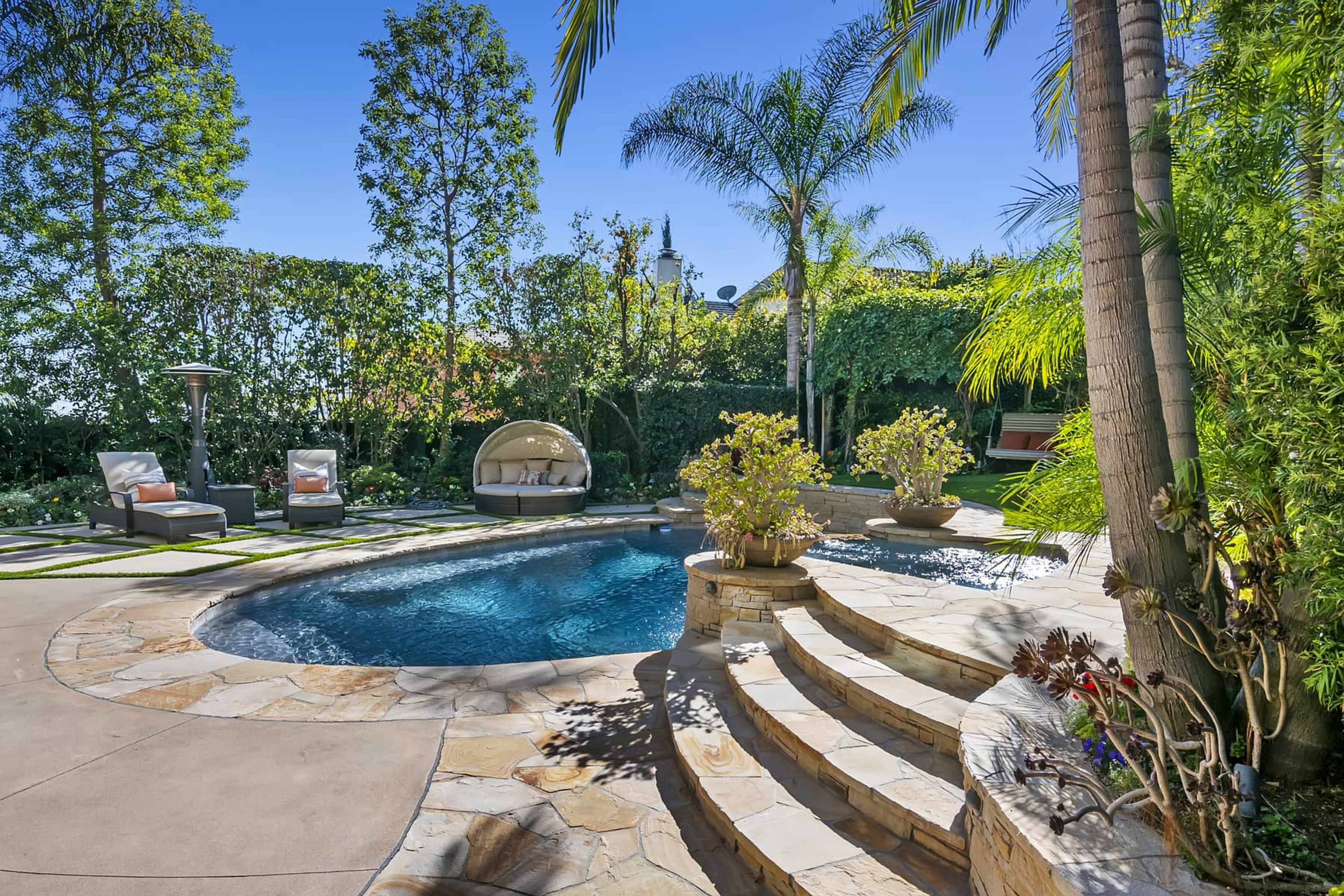 The image shows a landscaped backyard featuring a pool surrounded by stone steps, lounge chairs, and tropical plants.