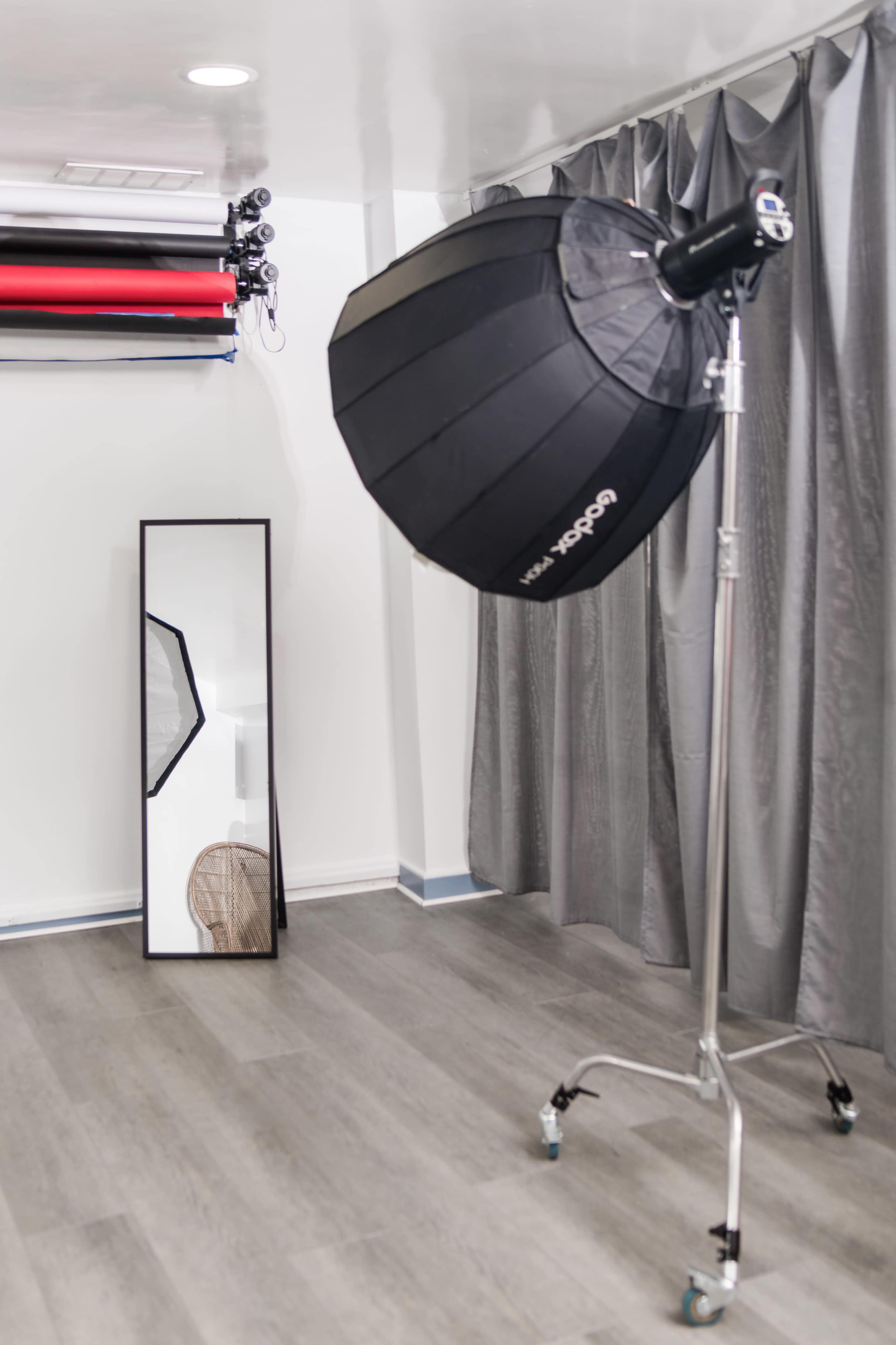 A photo studio setup with a large softbox light on a stand, a full-length mirror, and colored backdrop rolls hanging on the wall.