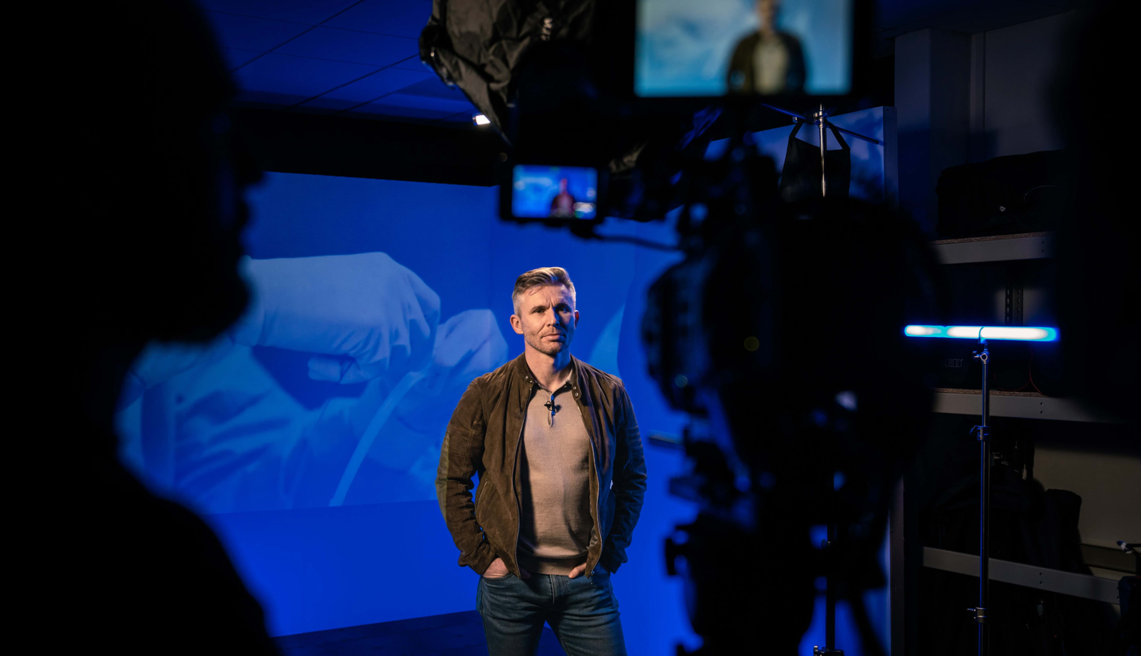 A man stands in front of a blue backdrop while being filmed, with silhouettes of camera equipment and crew visible in the foreground.