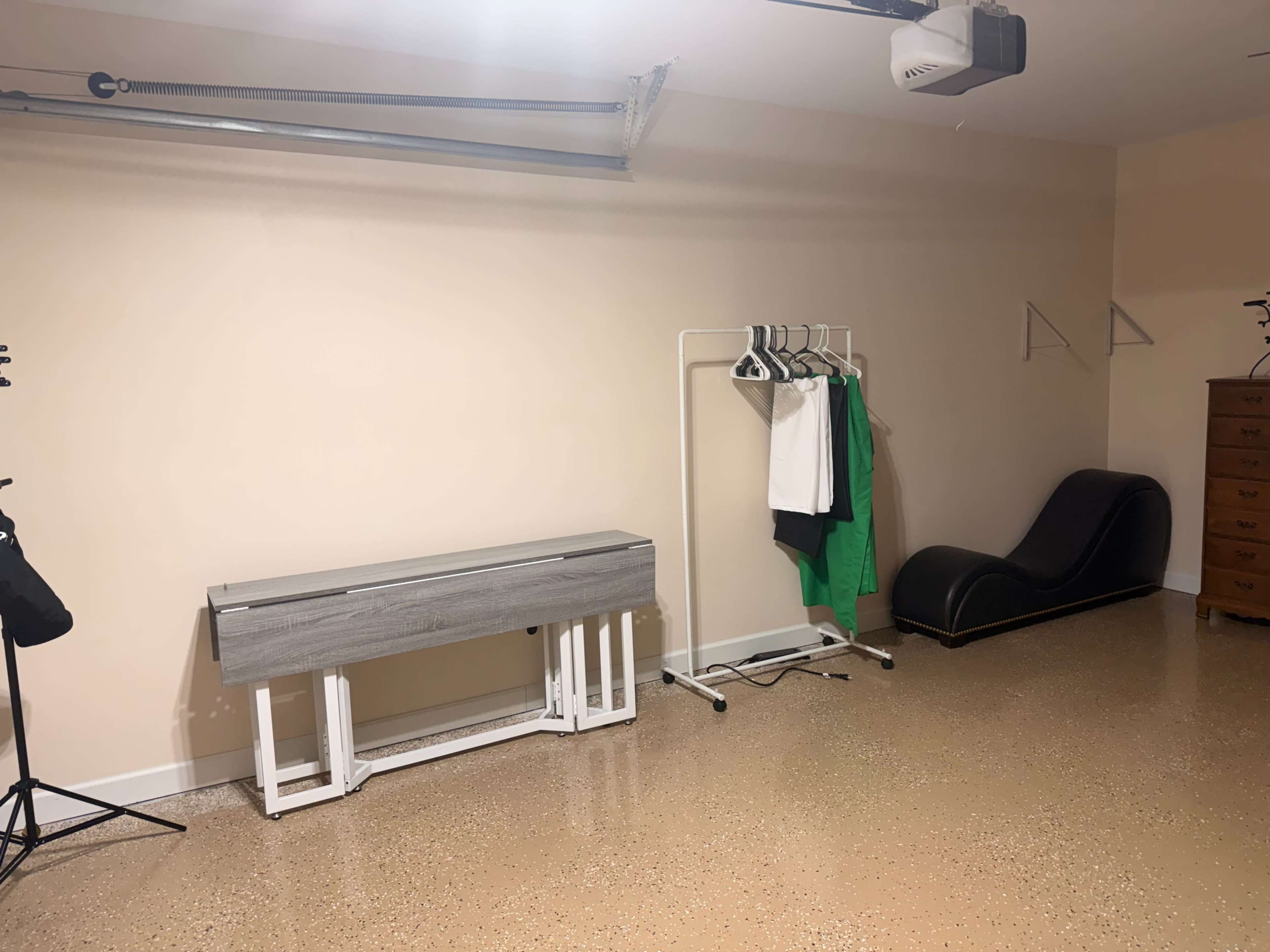 The image shows a garage with a light-colored wall, a storage bench, a clothing rack with garments, and a lounge chair.
