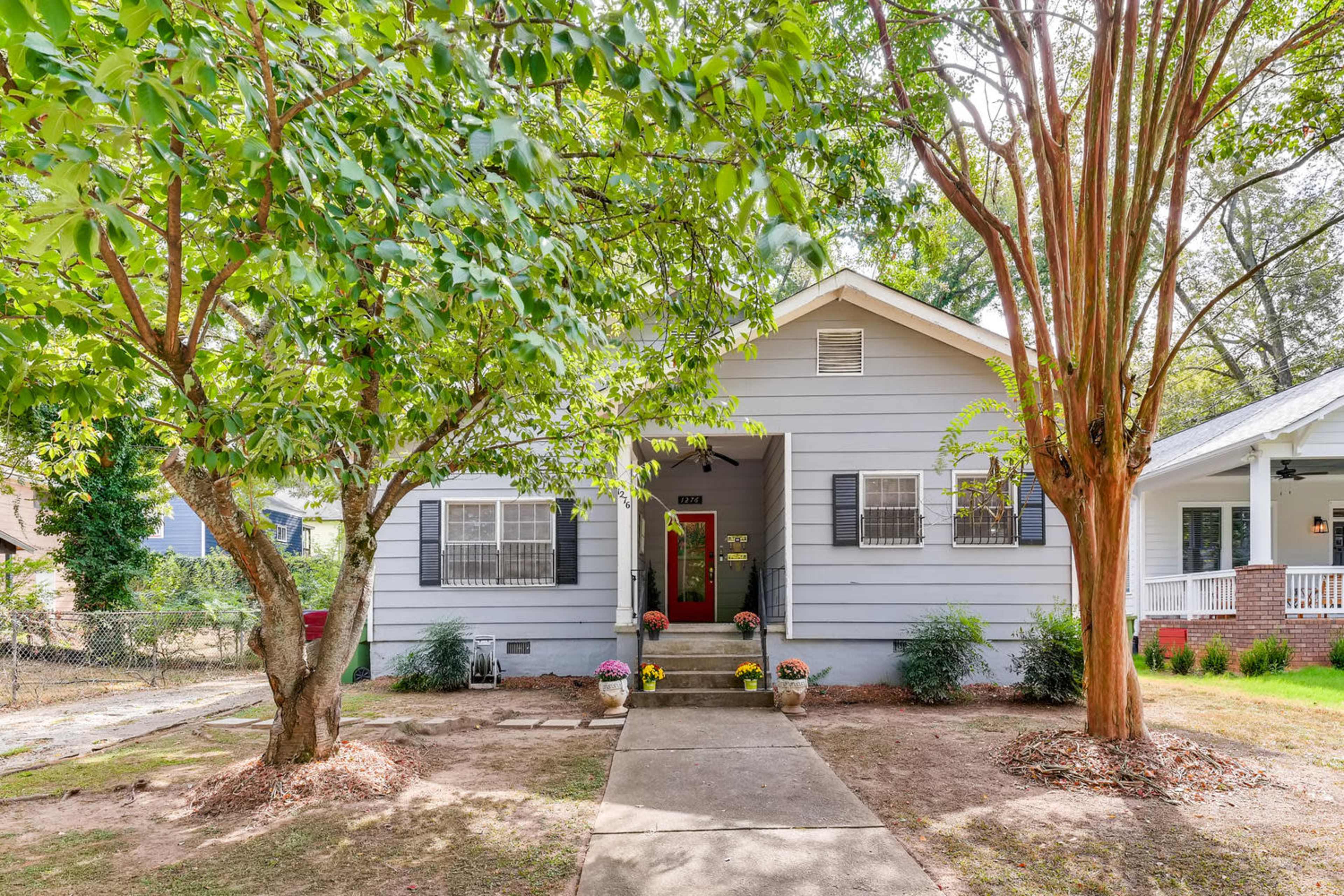 A single-story gray house with a red front door is surrounded by trees and flower pots in the front yard.