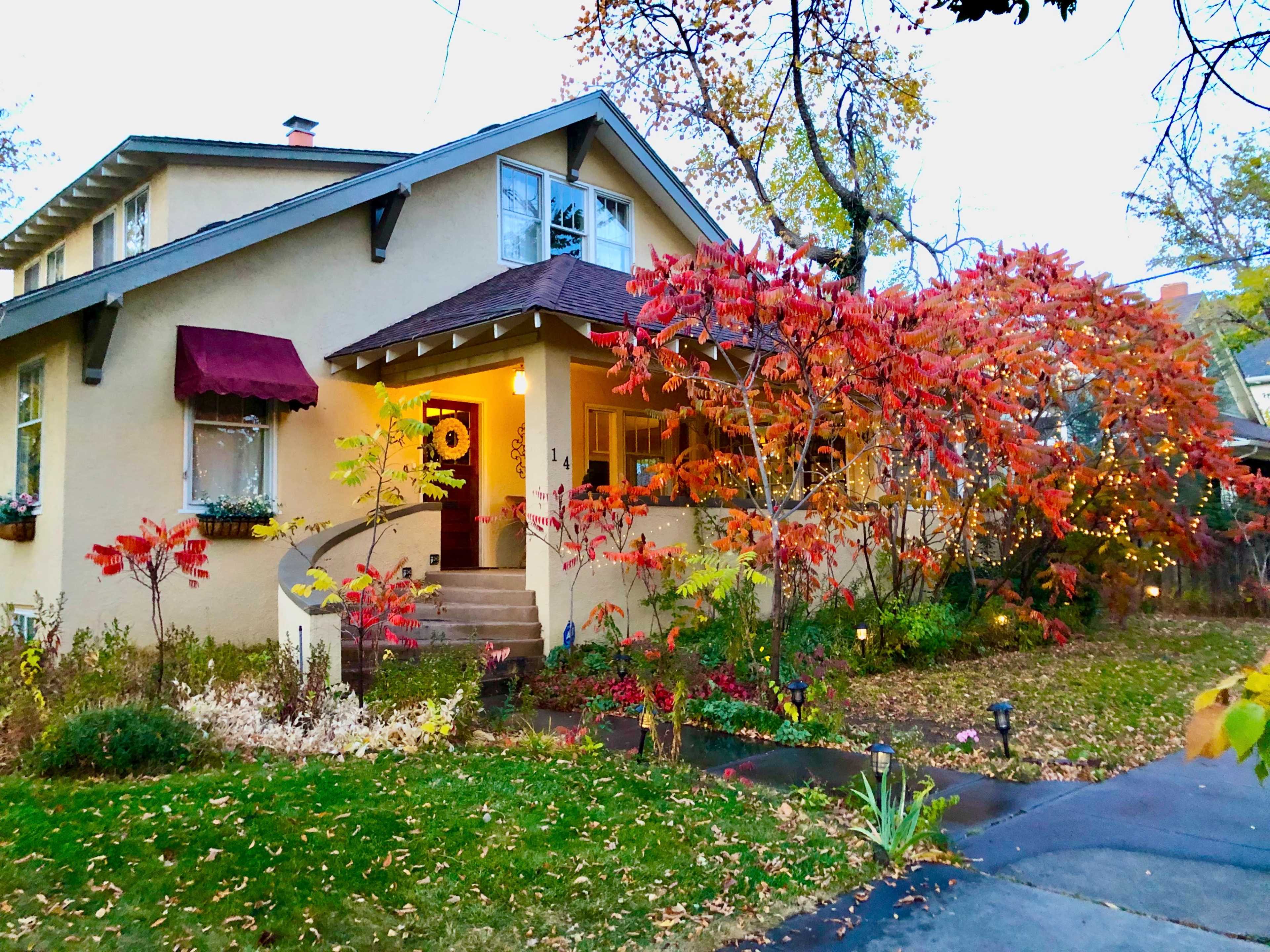 A two-story house with a maroon awning features colorful autumn foliage in the front yard and a well-kept garden.
