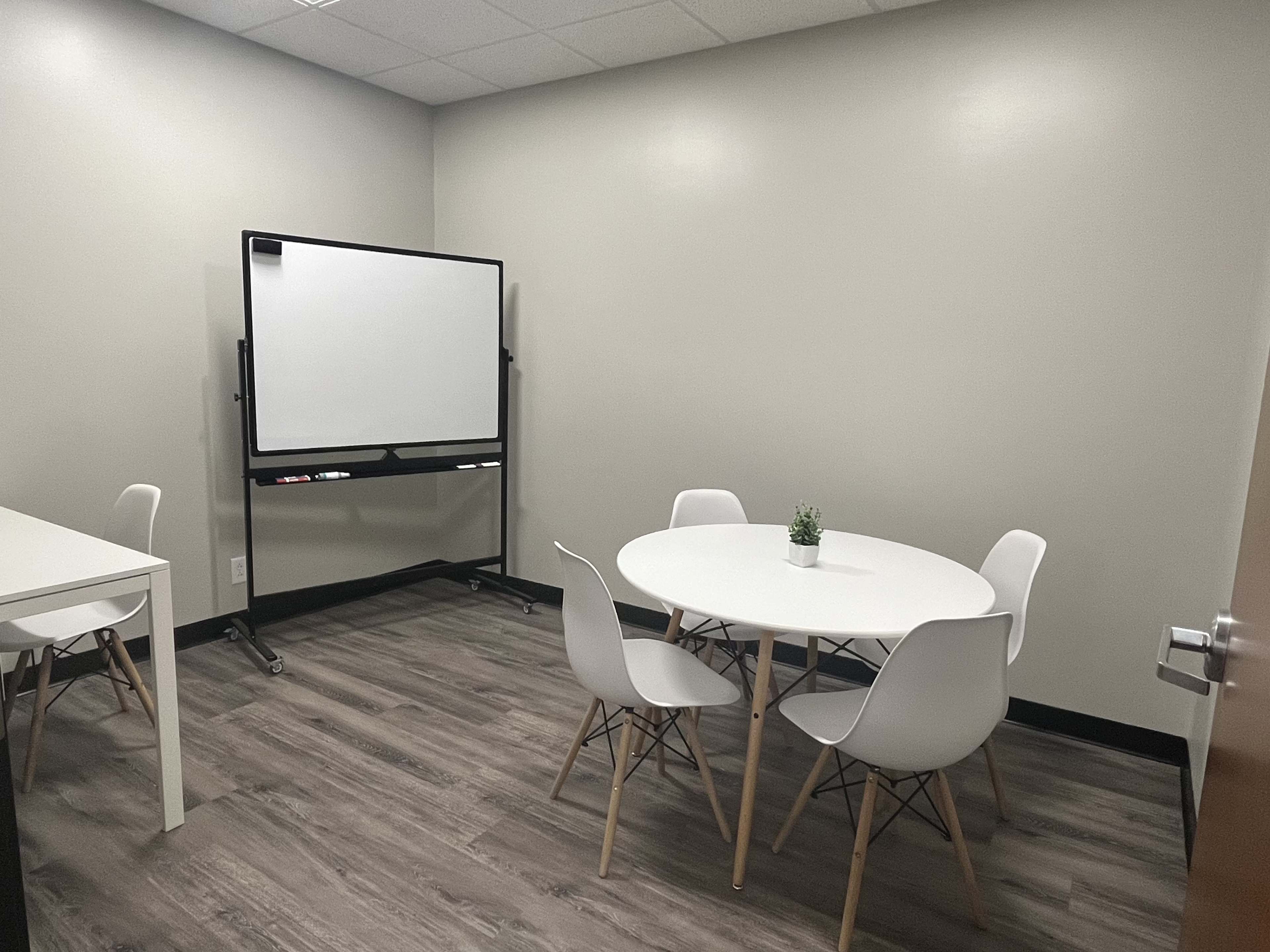 A small meeting room with a round table and four chairs, a whiteboard on wheels, and a minimalistic design.