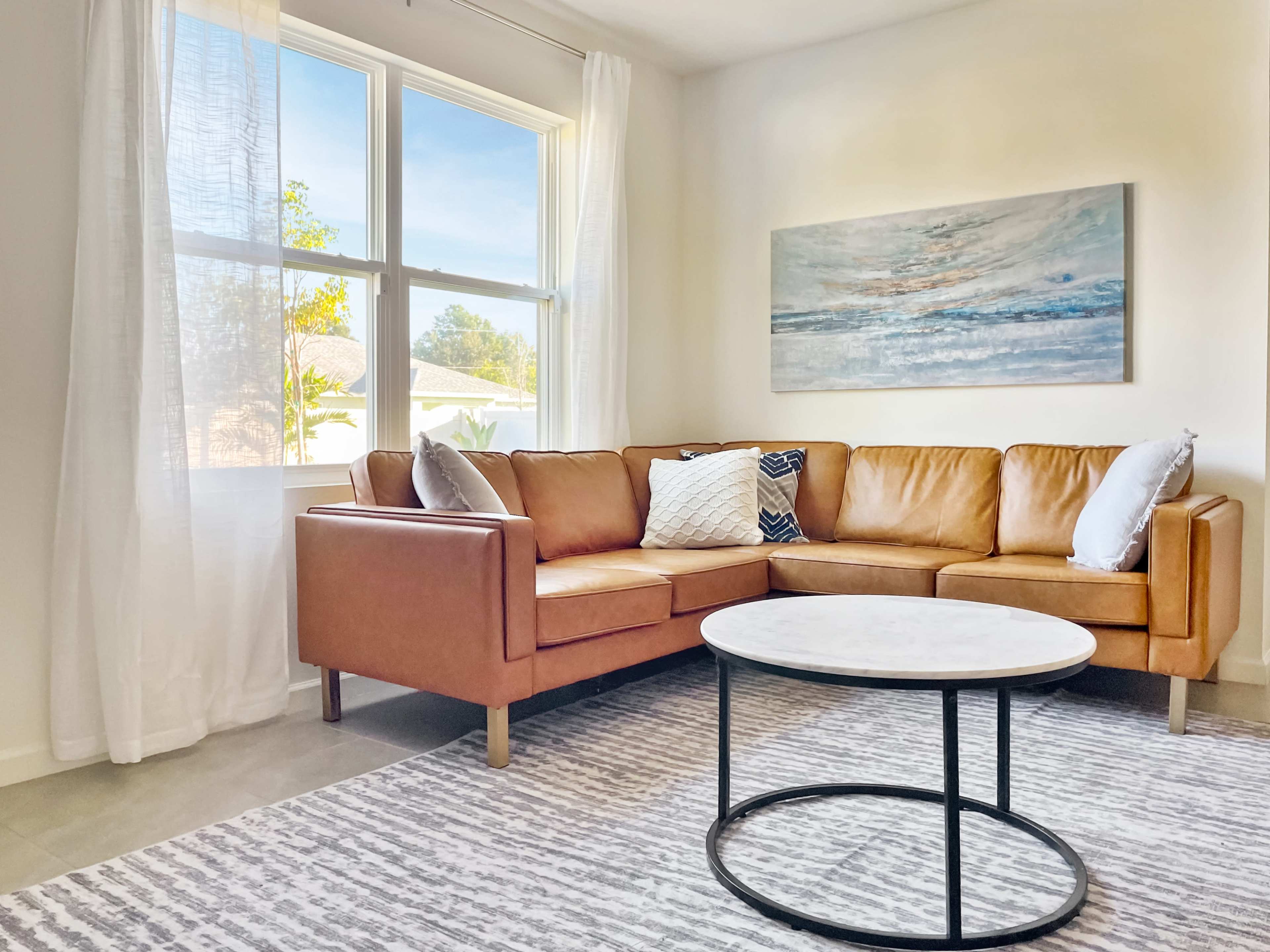 A modern living room with a tan leather sectional sofa, a round marble coffee table, and a large window allowing natural light to fill the space.