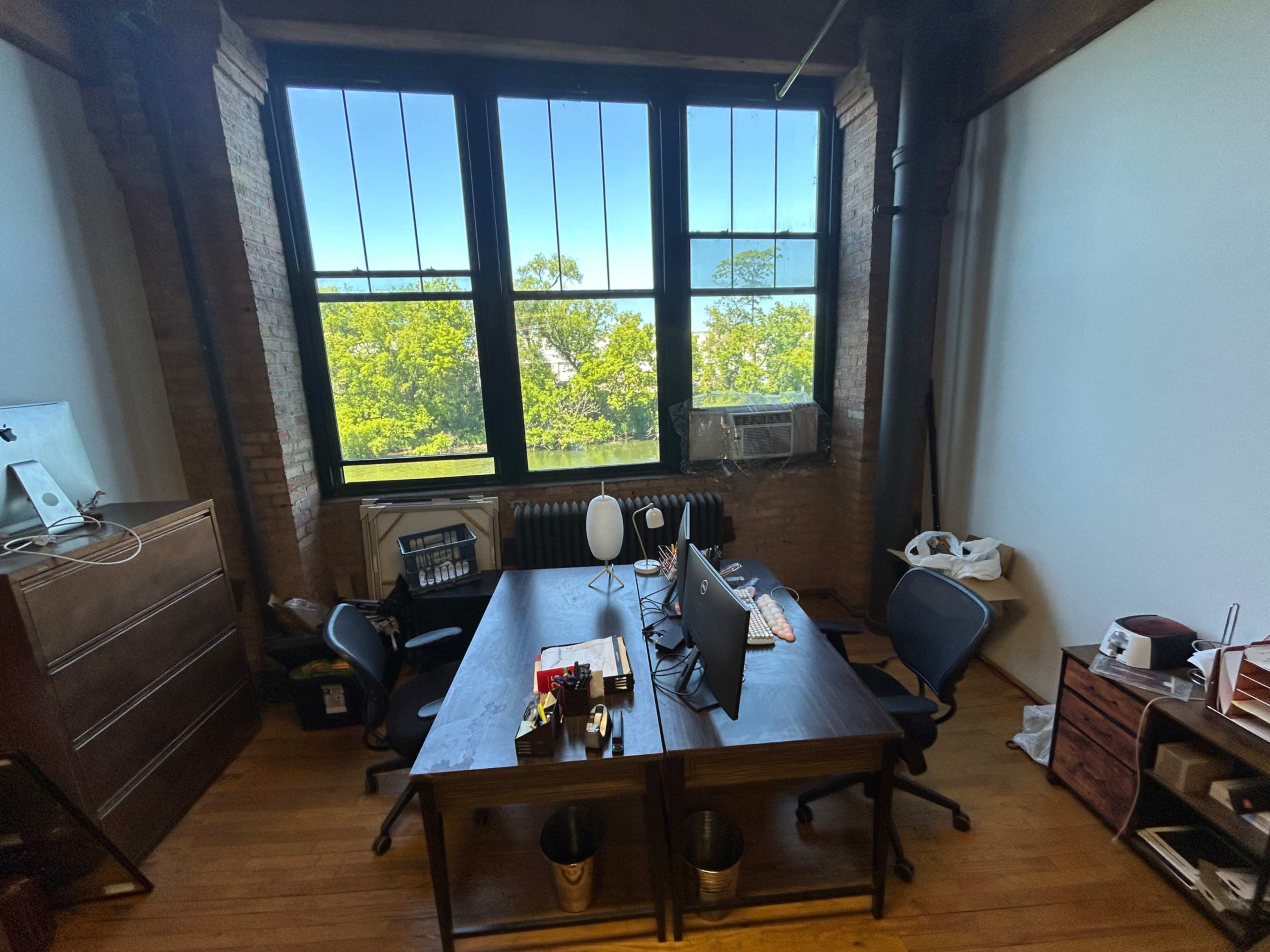 The image shows a spacious office with two desks facing each other and large windows that overlook greenery outside.