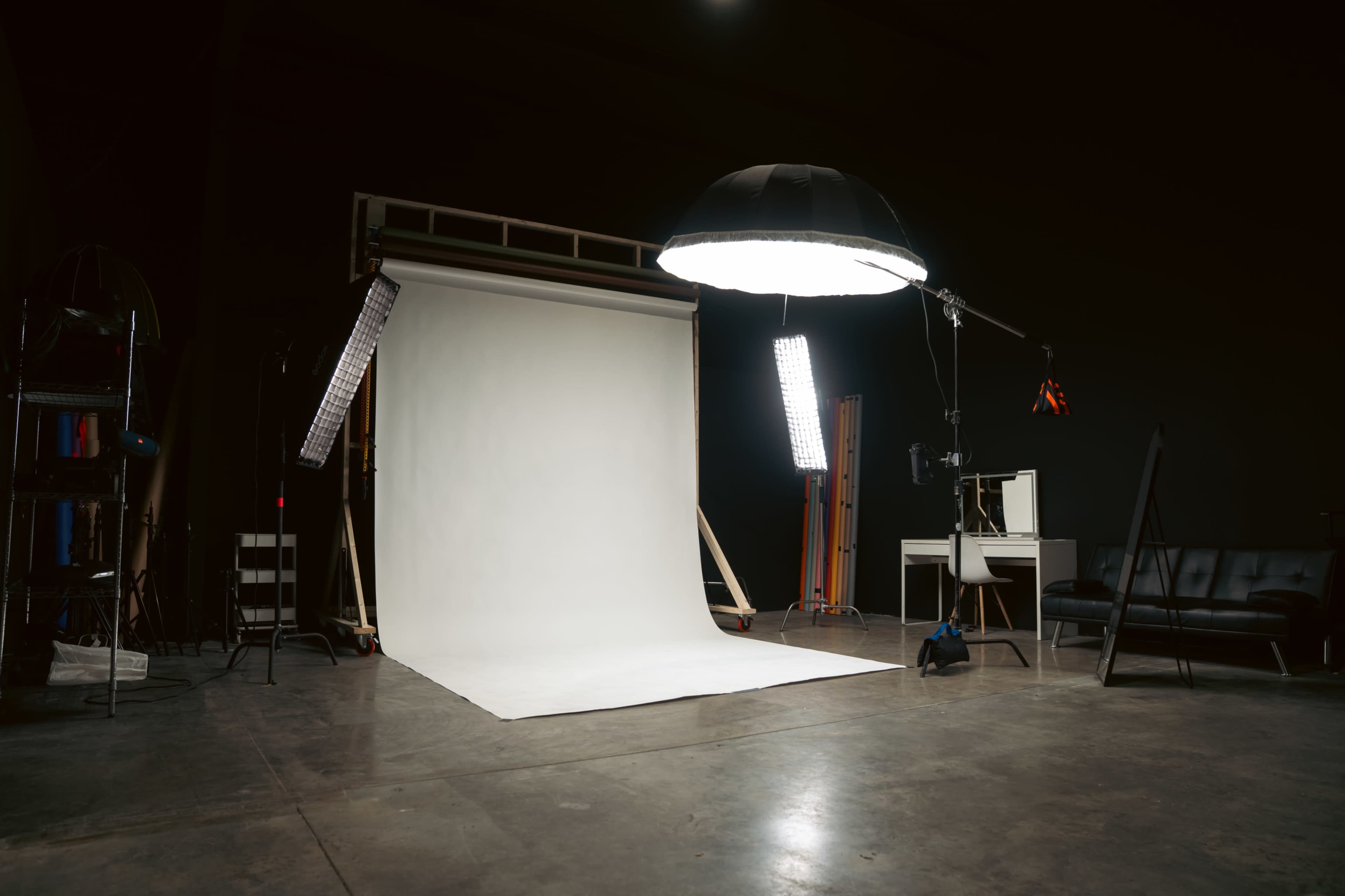 The image shows a photography studio with a white backdrop, softbox lights, and a black leather couch in a dimly lit space.