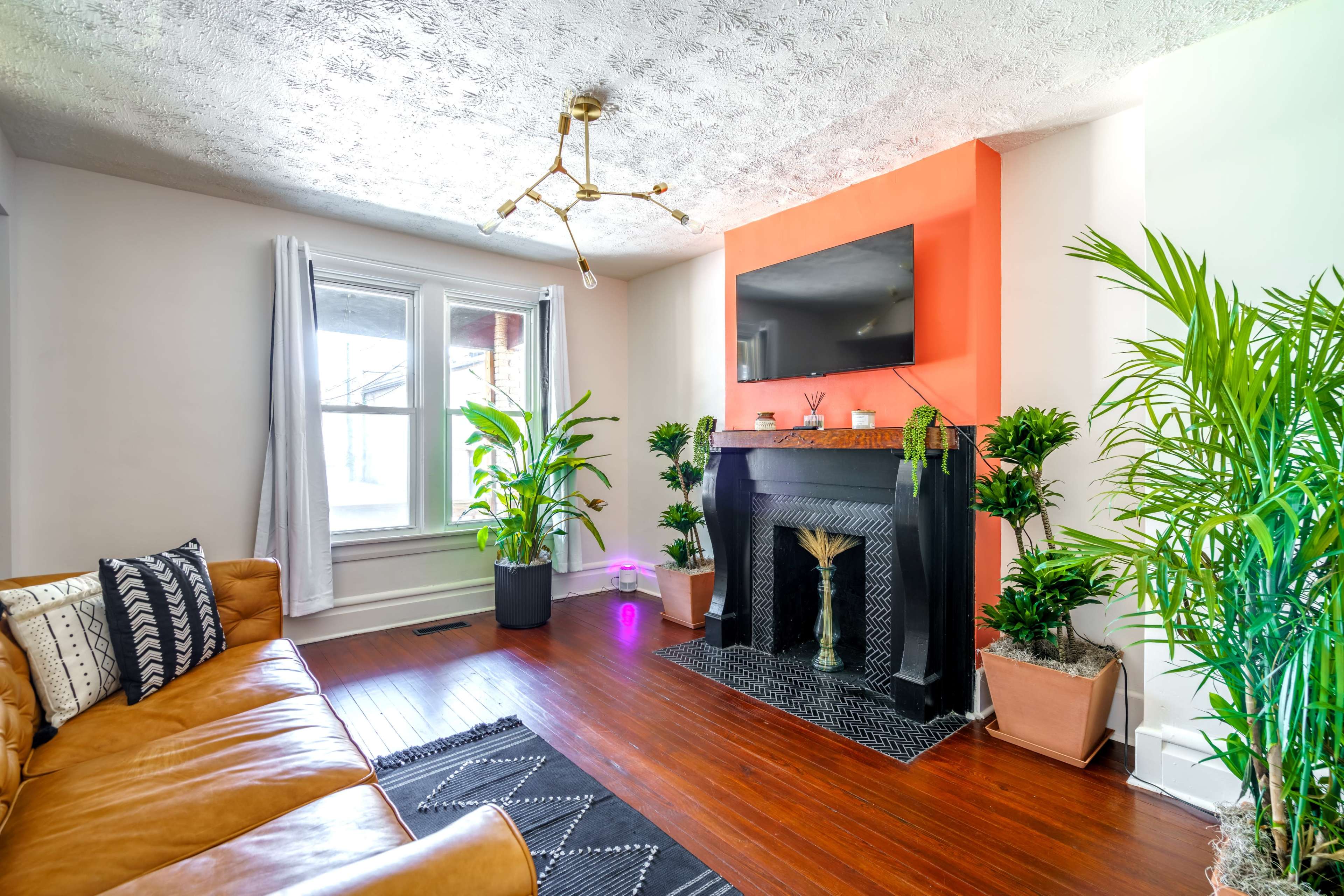 The image shows a cozy living room featuring a tan leather sofa, a black and white rug, a decorative fireplace, potted plants, and a mounted television on an orange accent wall.
