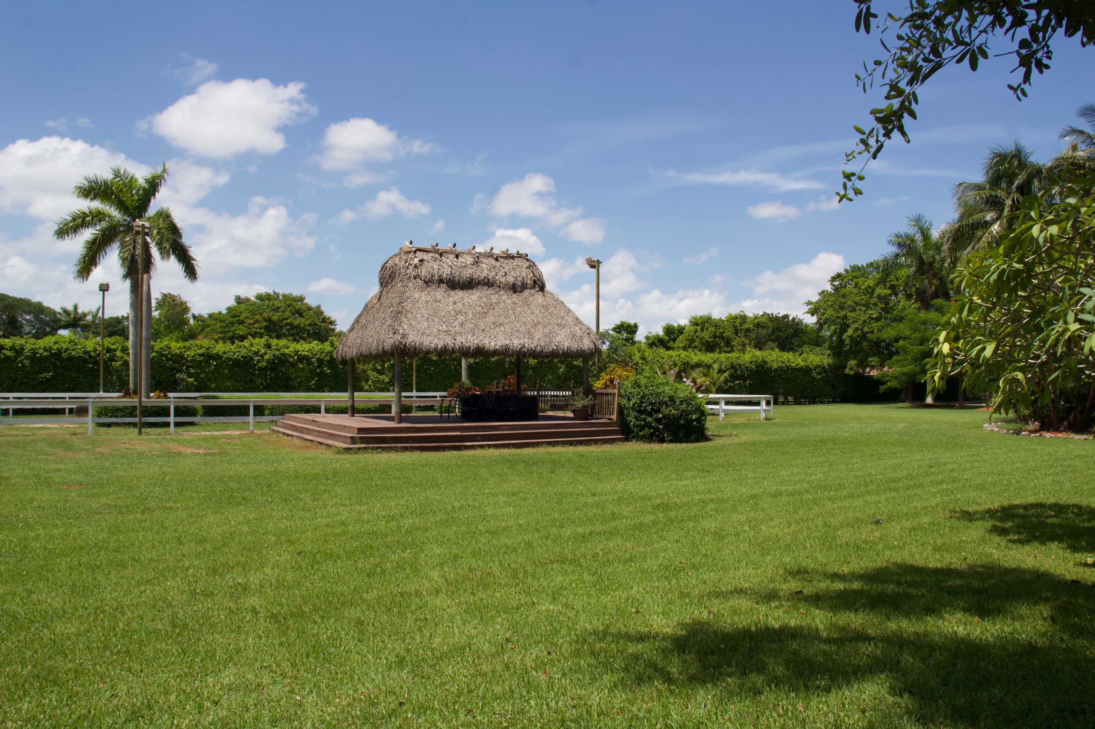 A thatched gazebo sits in a well-maintained lawn surrounded by lush greenery and palm trees under a partly cloudy sky.