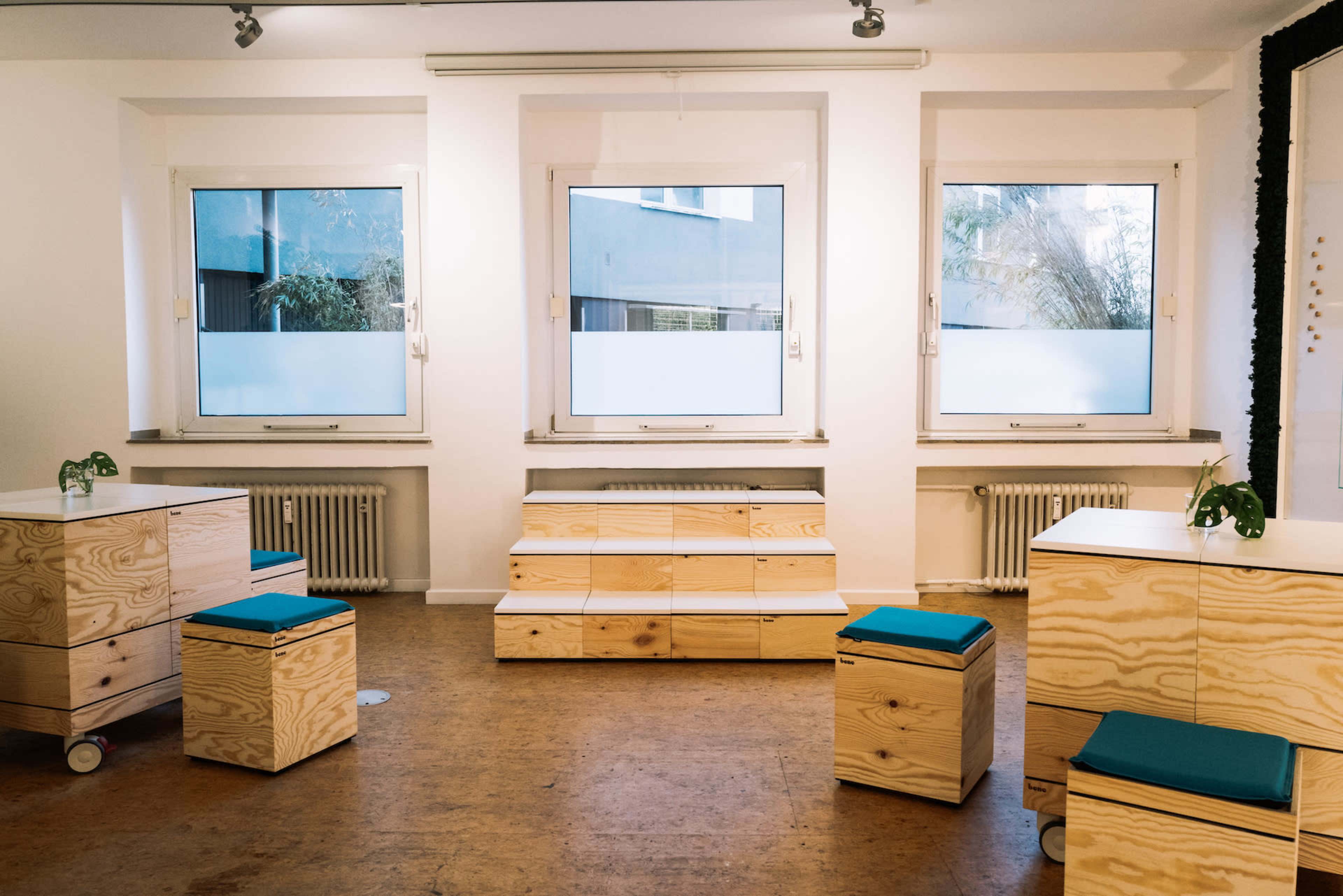 The image shows a bright room with three windows, featuring minimalist wooden furniture including stacked boxes and seats with blue cushions.