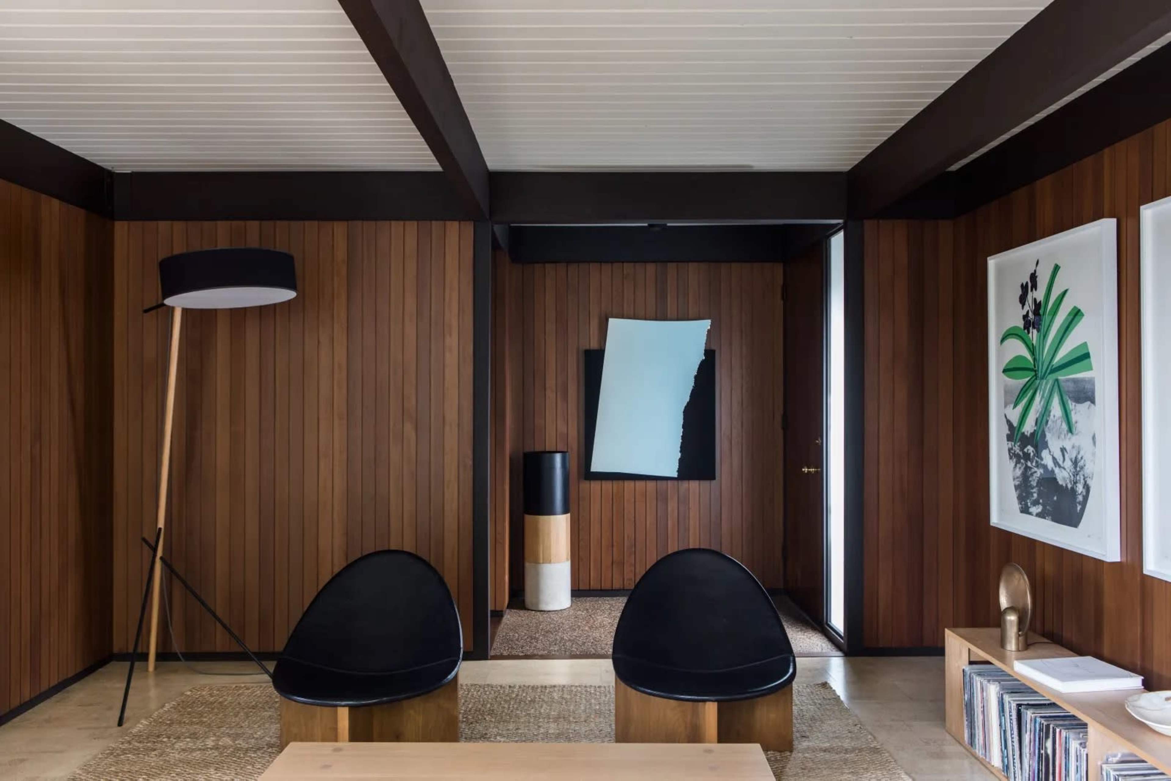 The interior features a modern living space with wooden paneling, two black chairs, a minimalist floor lamp, and art displayed on the walls.