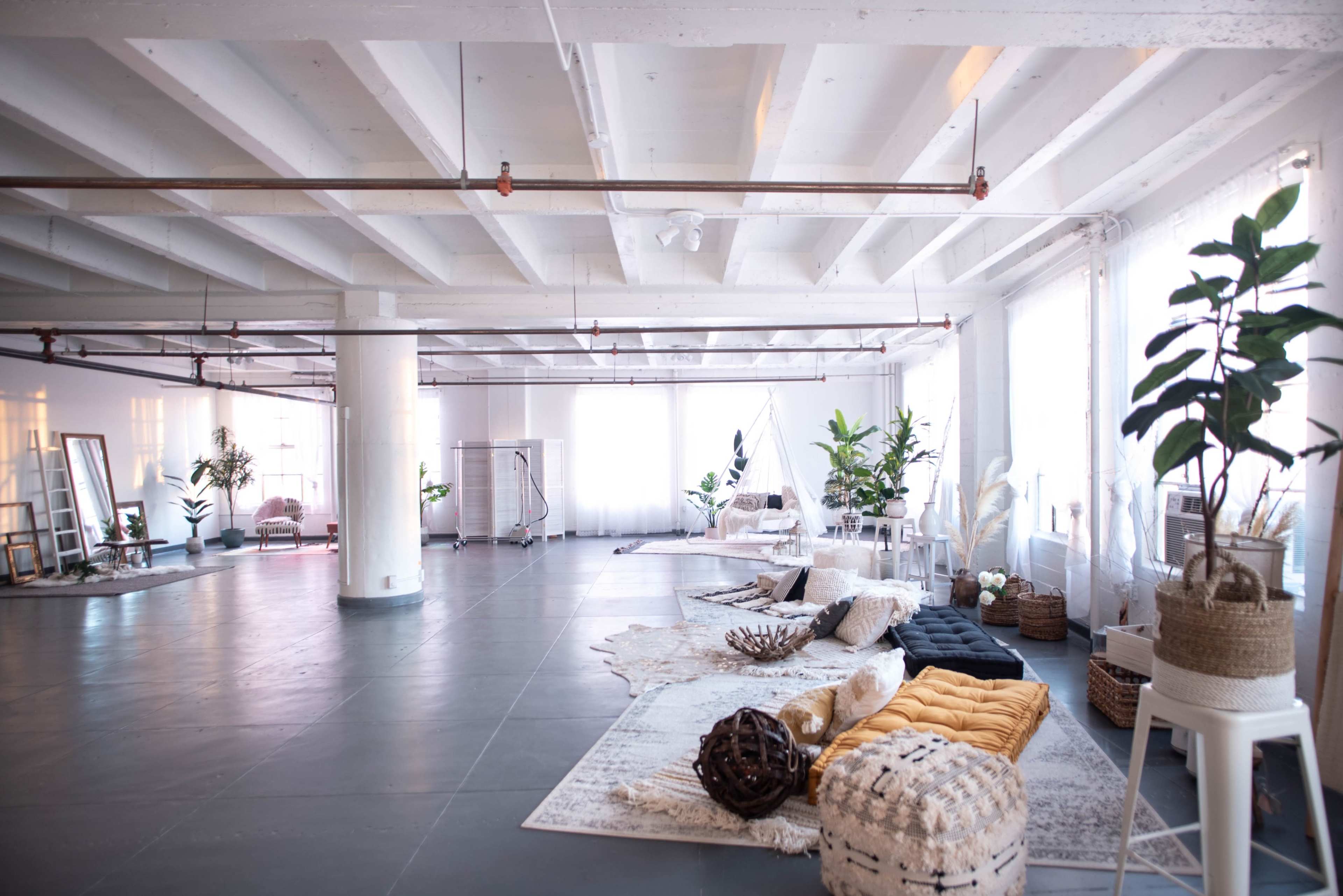 A spacious, minimalist studio filled with various seating areas, plants, and textile decor under bright natural light.