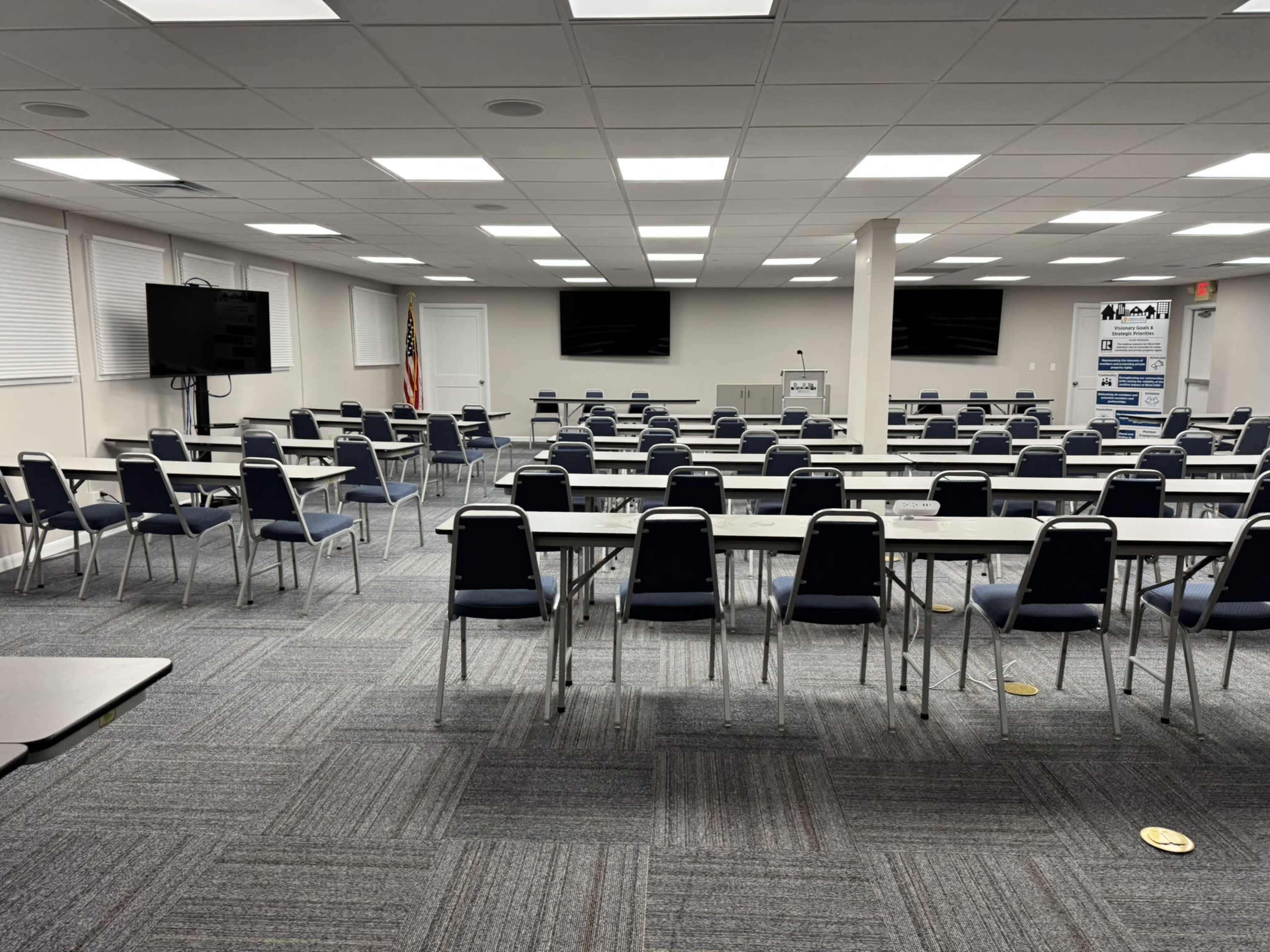 The image shows a large meeting room arranged with multiple rows of tables and chairs, equipped with audiovisual equipment and a podium.
