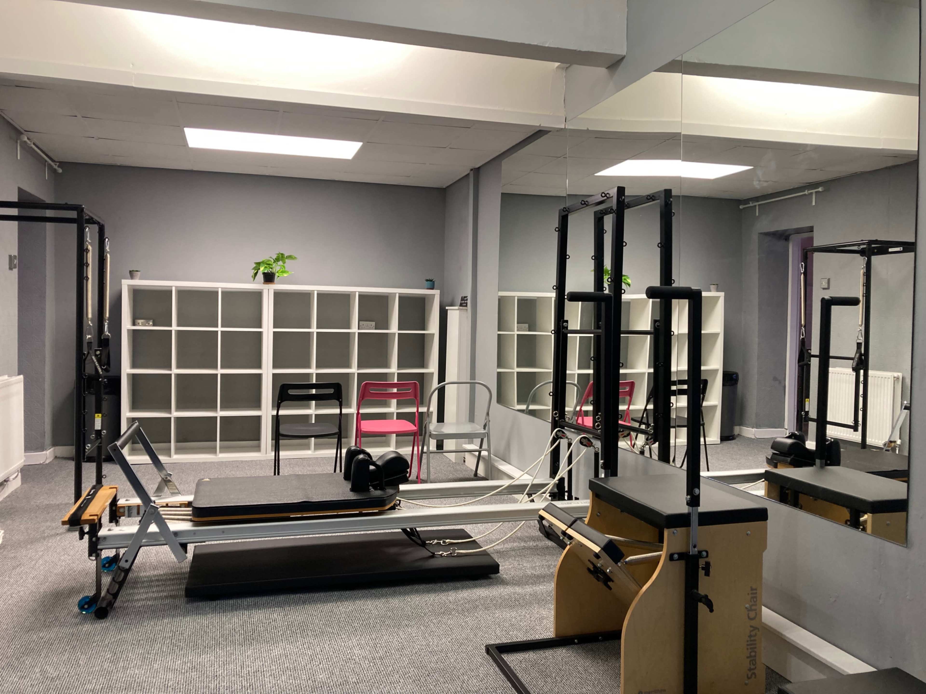 A fitness studio equipped with a reformer machine, exercise equipment, and a shelf of chairs against a gray wall.