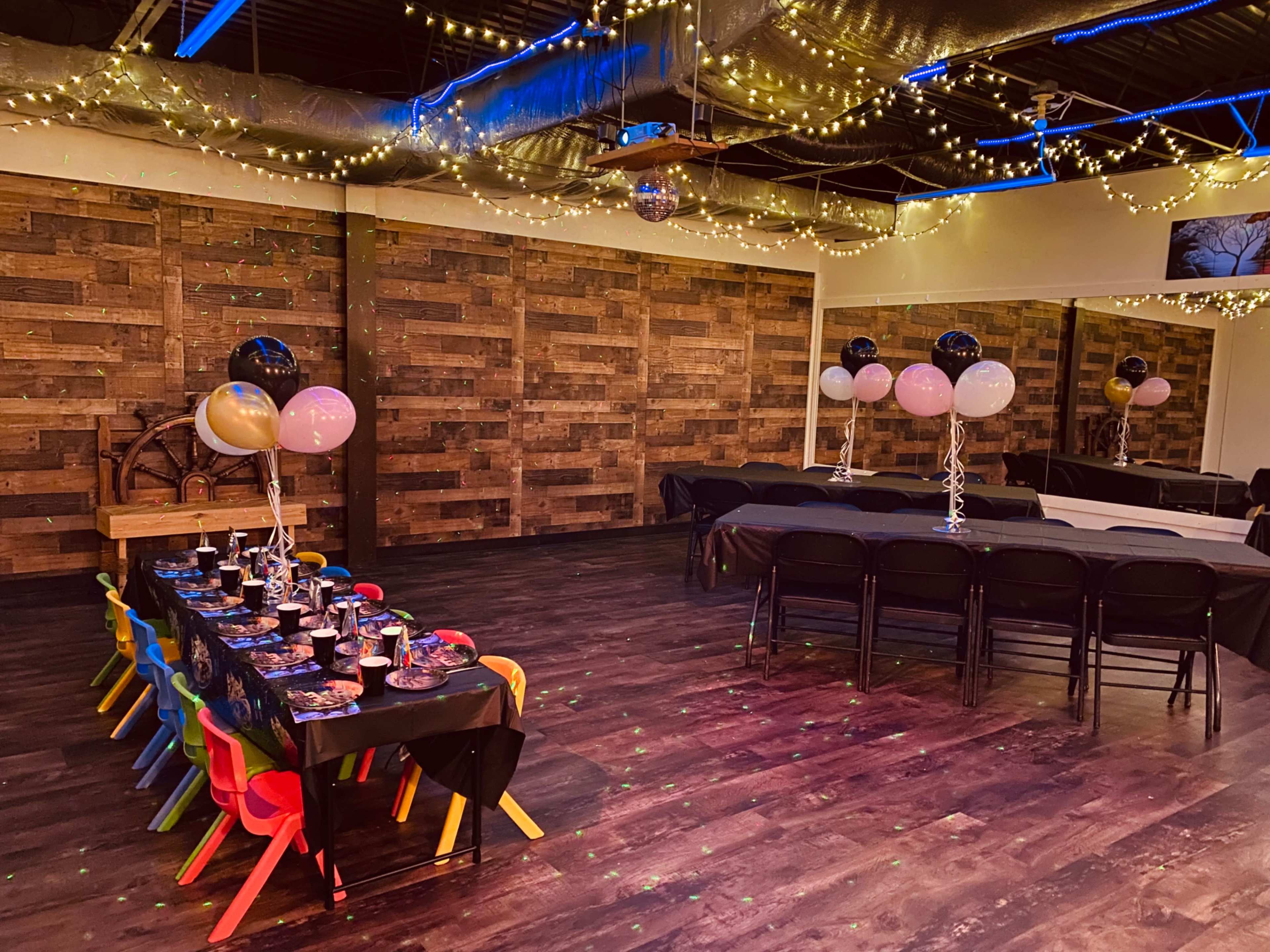 The image shows a decorated event space with colorful chairs at a table set for a celebration, balloons, and string lights hanging from the ceiling.