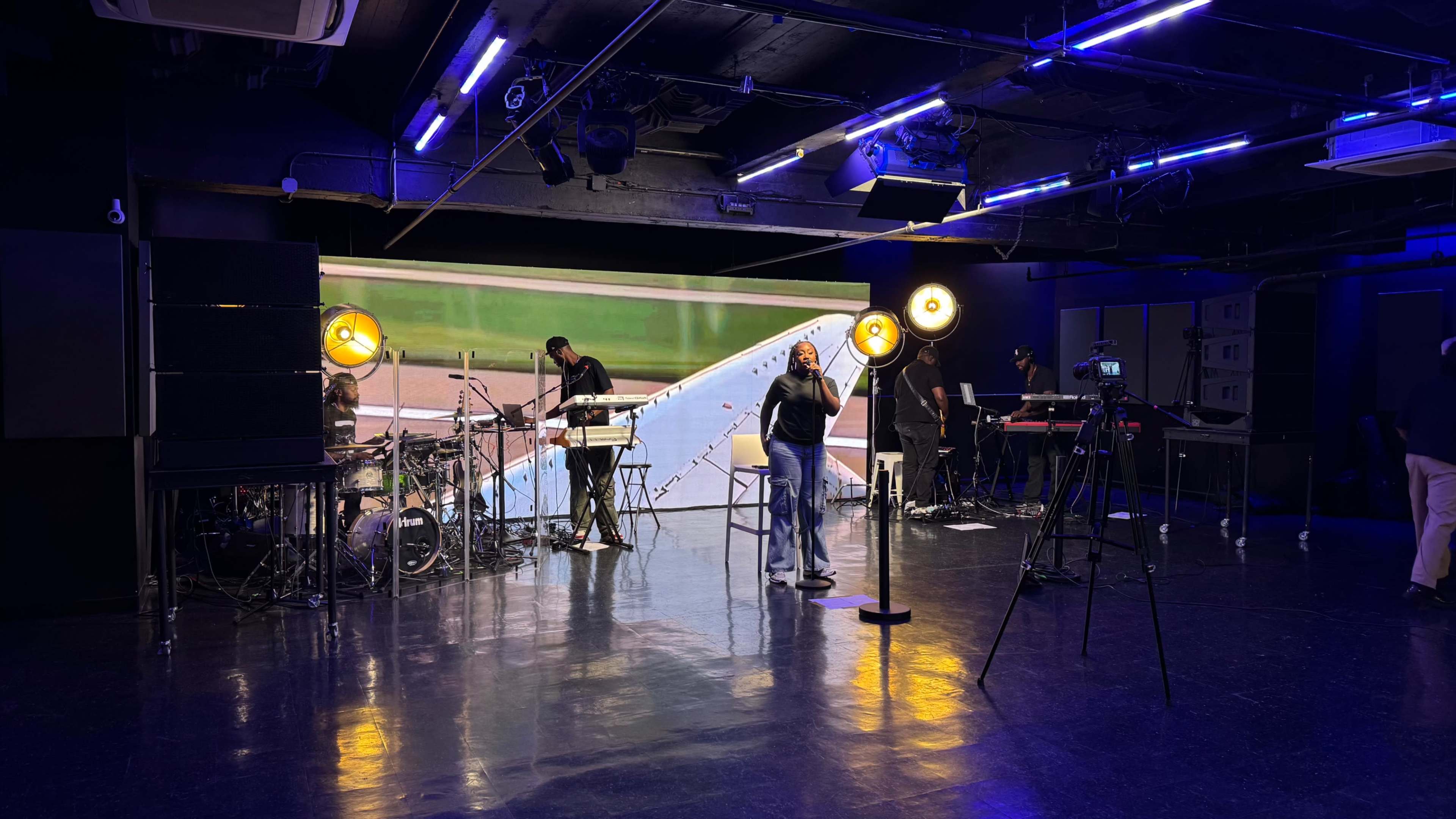 A music setup is being arranged in a studio with musicians and equipment in the background, while a performer stands in front of cameras and lights.