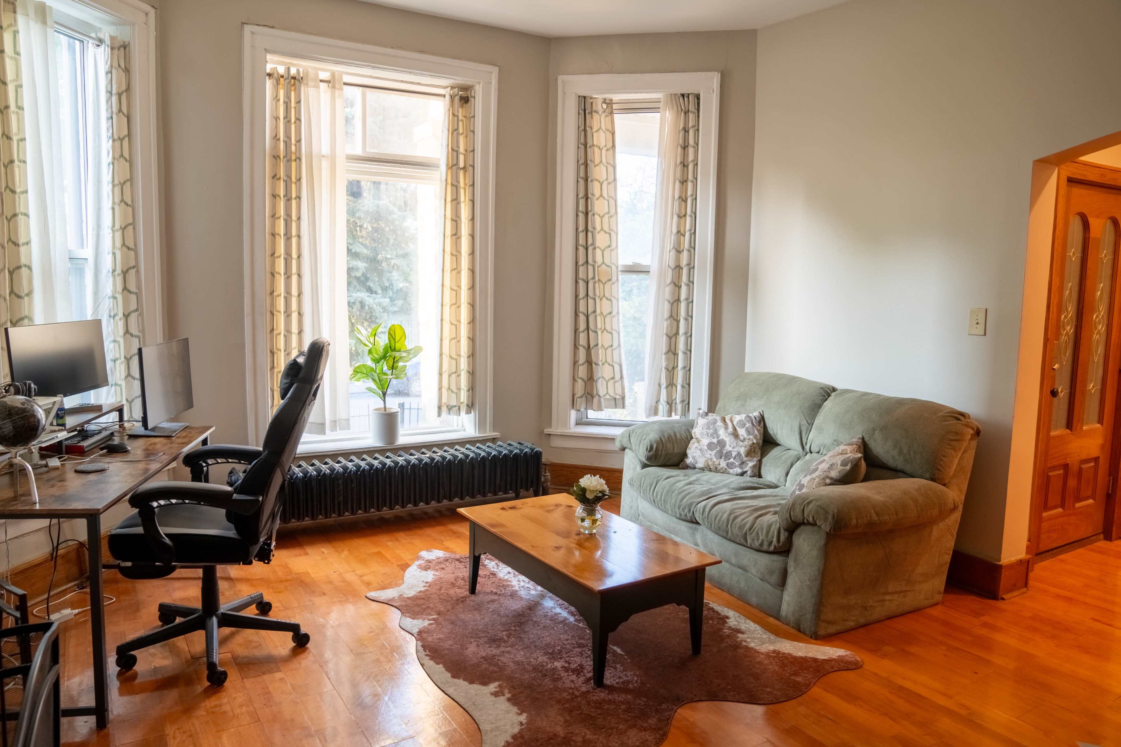 A well-lit living room with a green sofa, a wooden coffee table, a desk with a computer setup, and large windows adorned with curtains.