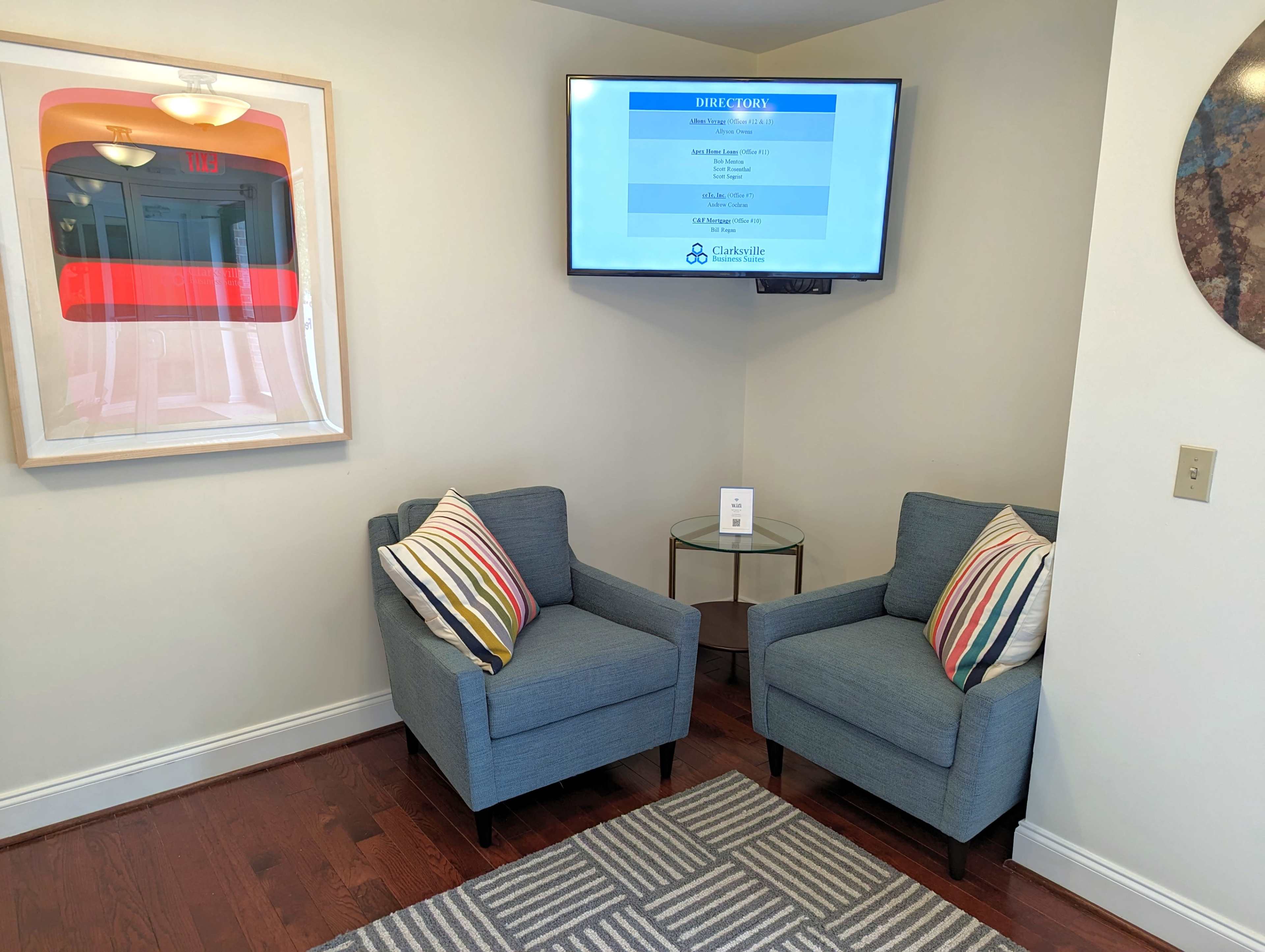 The image shows a small seating area with two blue chairs and a round table, accompanied by a mounted television displaying information on a wall.