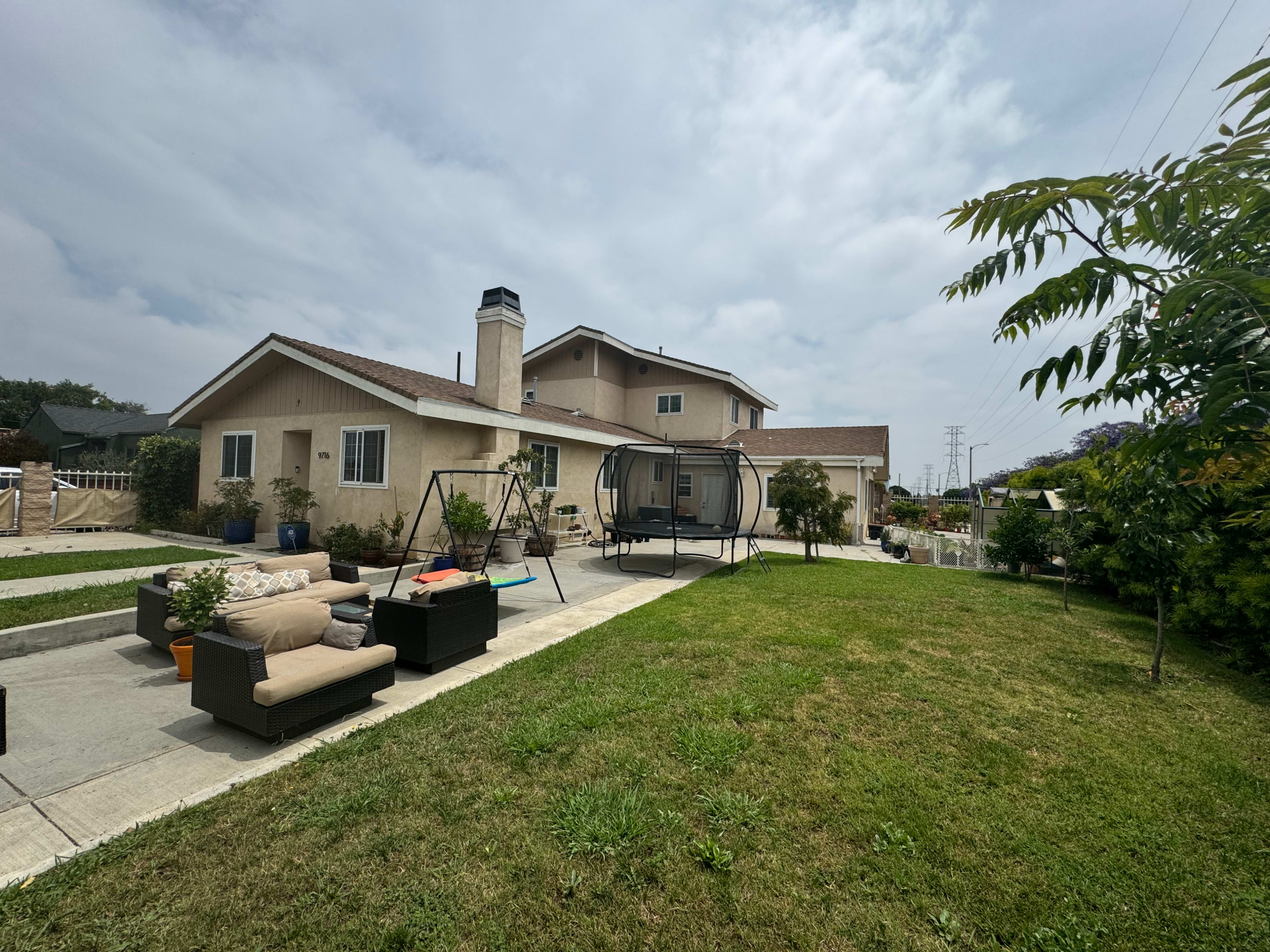 The image shows a backyard with a patio area featuring outdoor furniture, a swing, and a well-maintained lawn beside a two-story house under a cloudy sky.