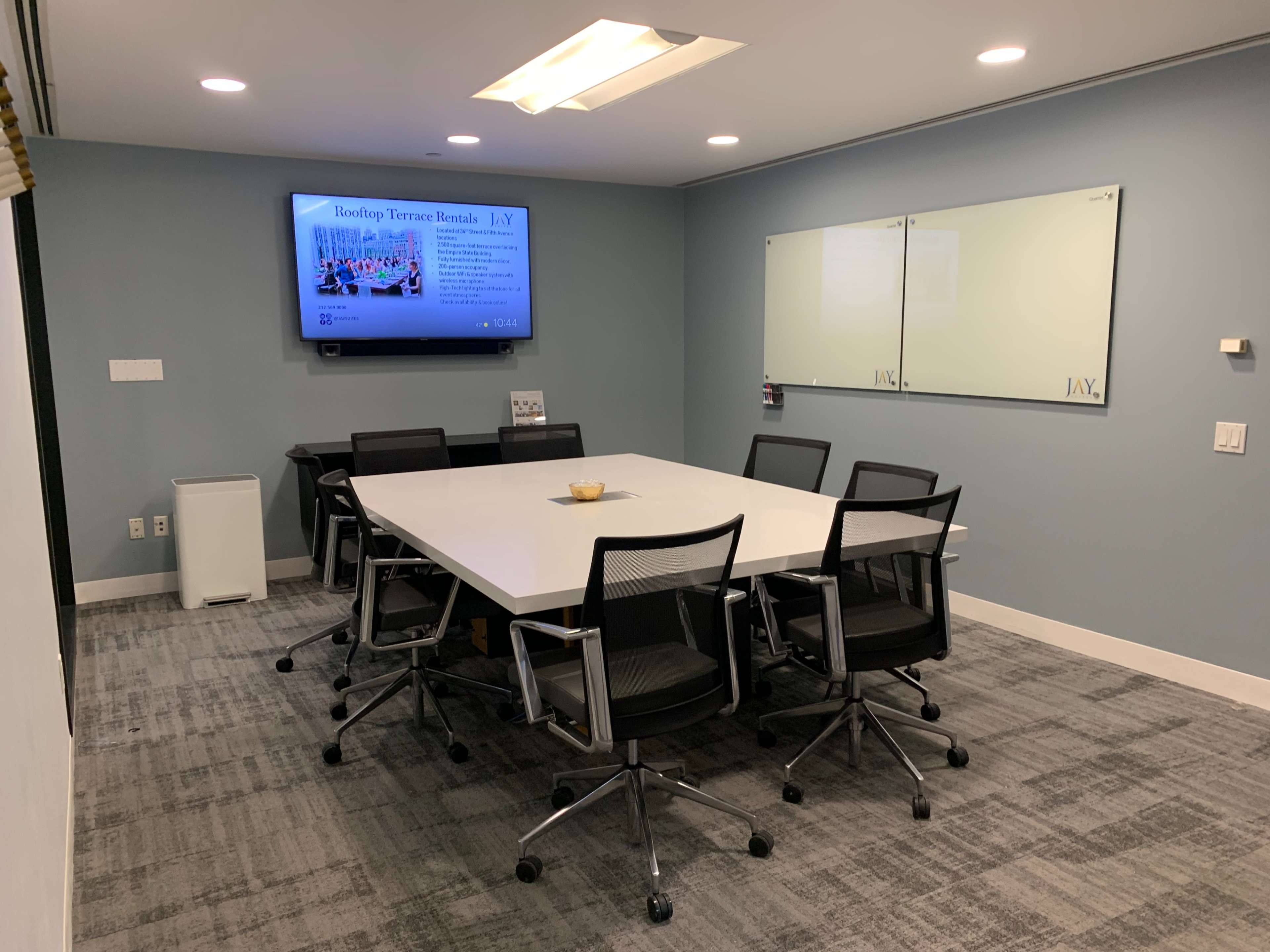 A modern conference room features a large white table surrounded by black chairs, with a wall-mounted screen displaying information, and a whiteboard on one side.