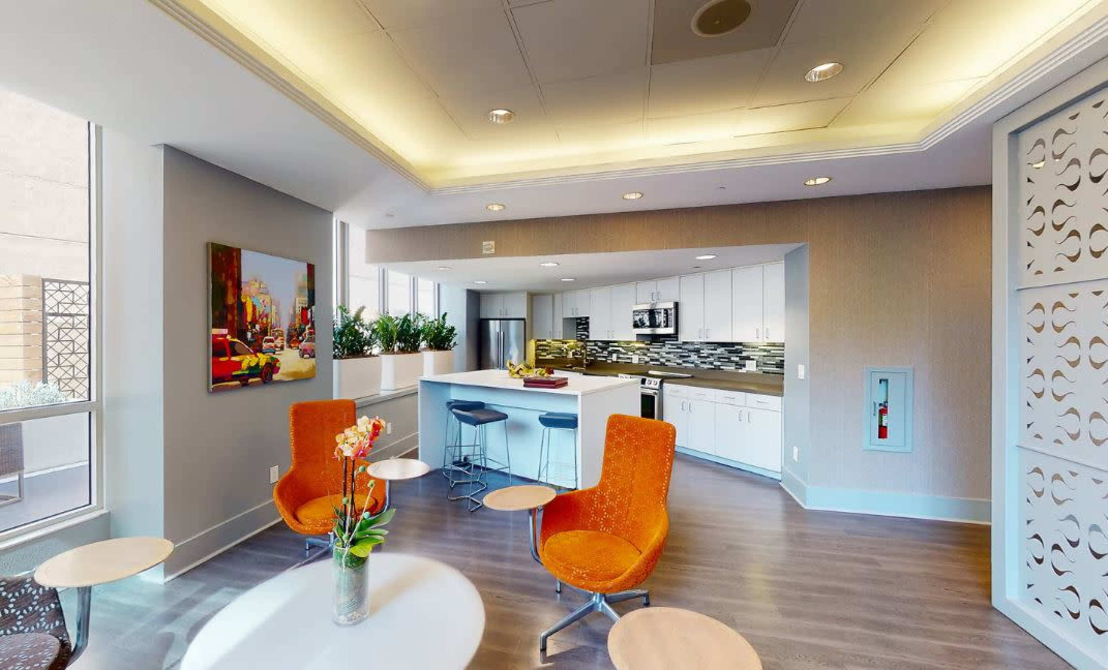 A modern kitchen and lounge area featuring orange chairs, a white countertop, and large windows with plants lining the interior.