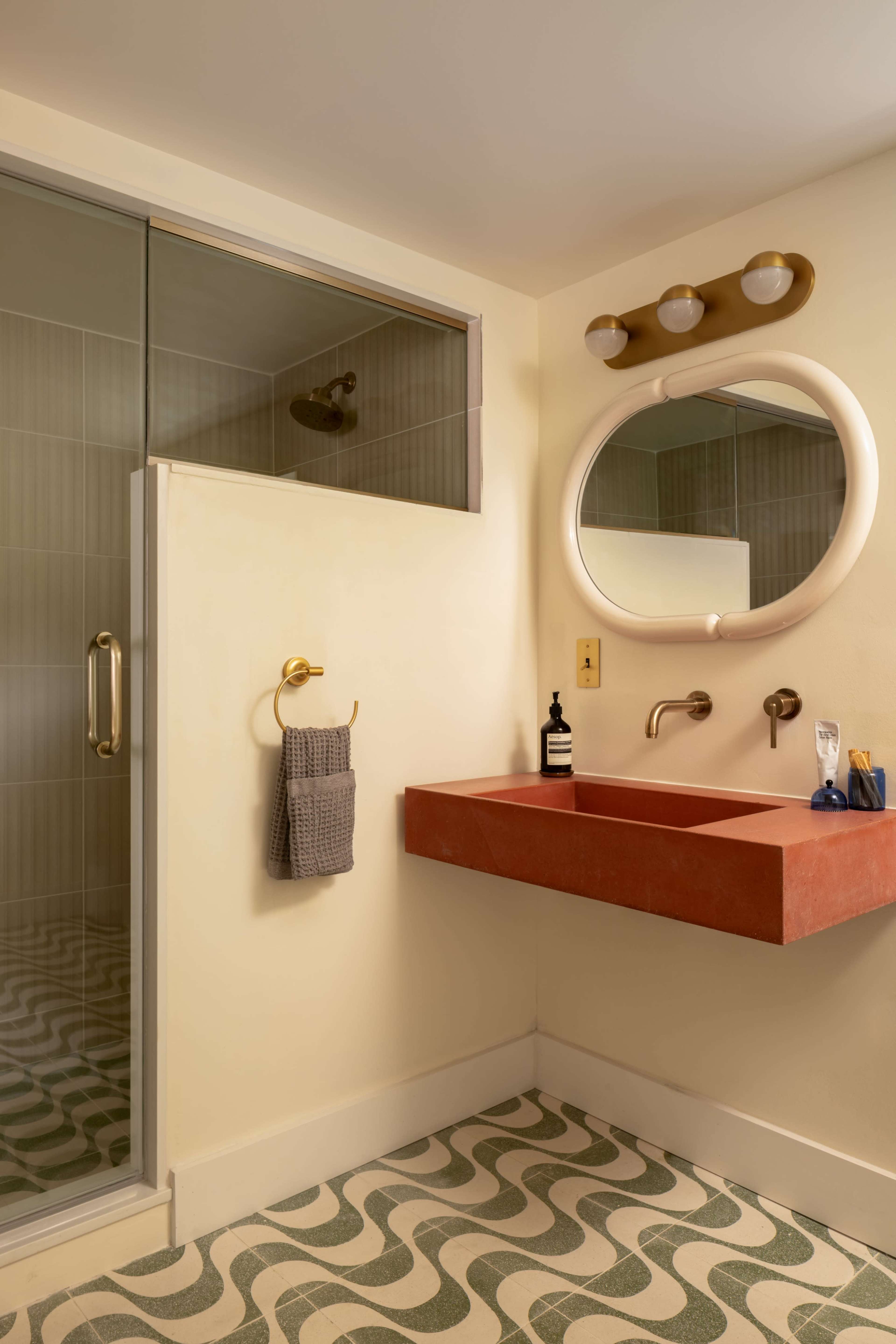The image shows a modern bathroom featuring a stone sink with two faucets, a circular mirror, a glass shower enclosure, and patterned flooring.