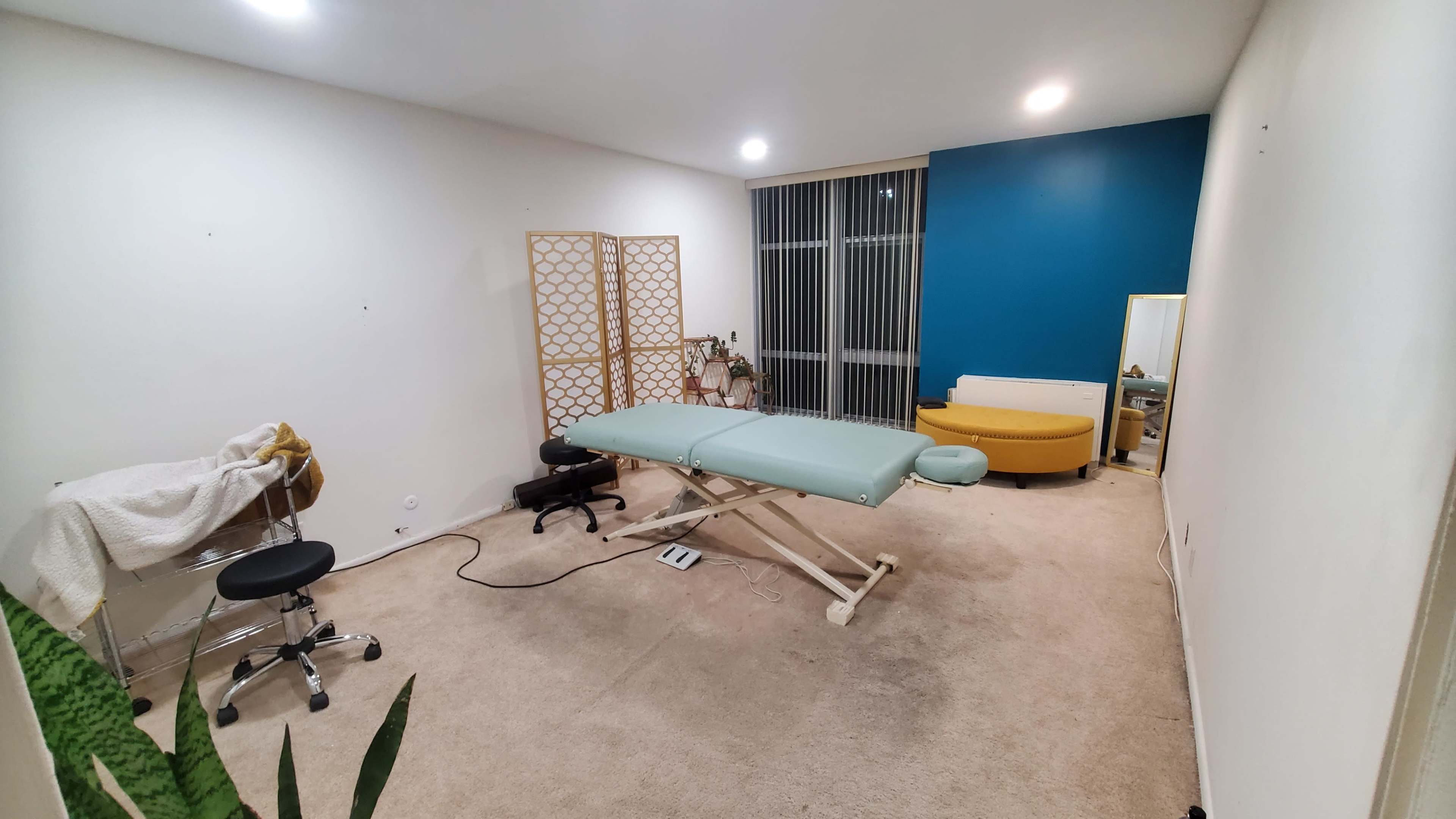 The image shows a spacious treatment room with a massage table, a privacy screen, a chair, and a yellow couch against a blue wall.