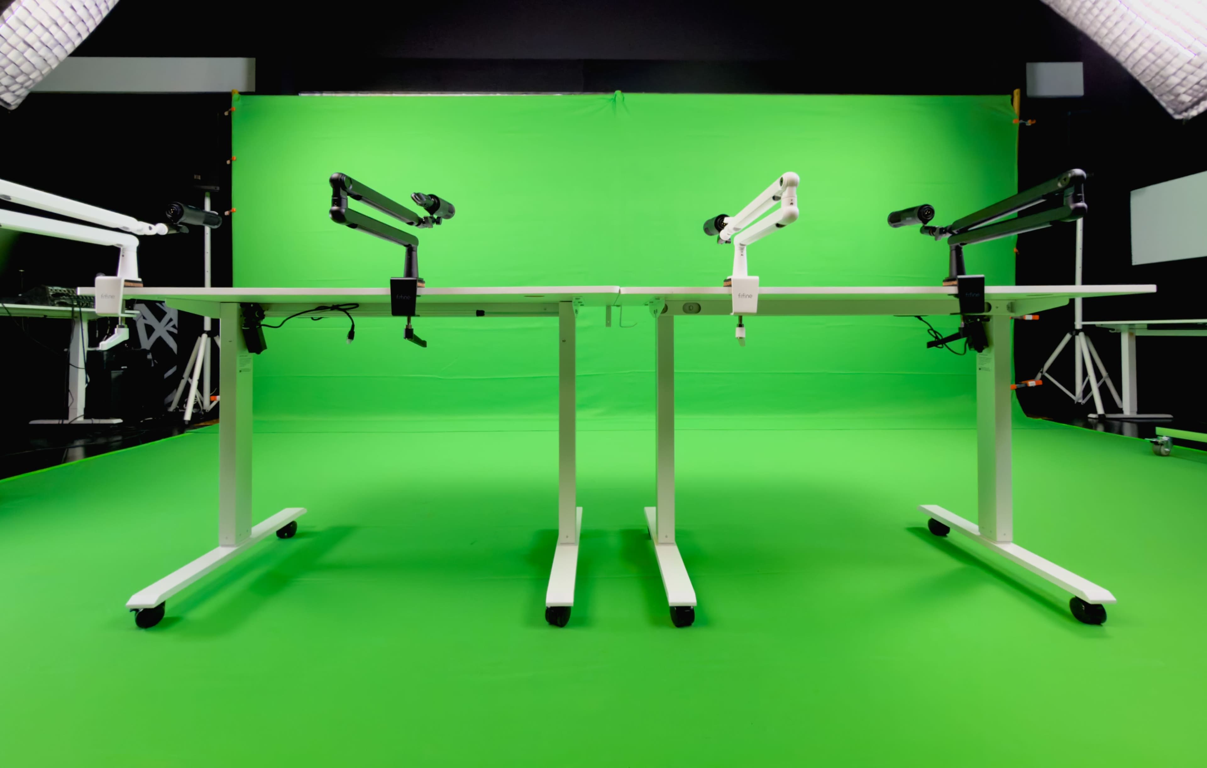 The image shows two adjustable desks with monitor arms positioned against a green backdrop.