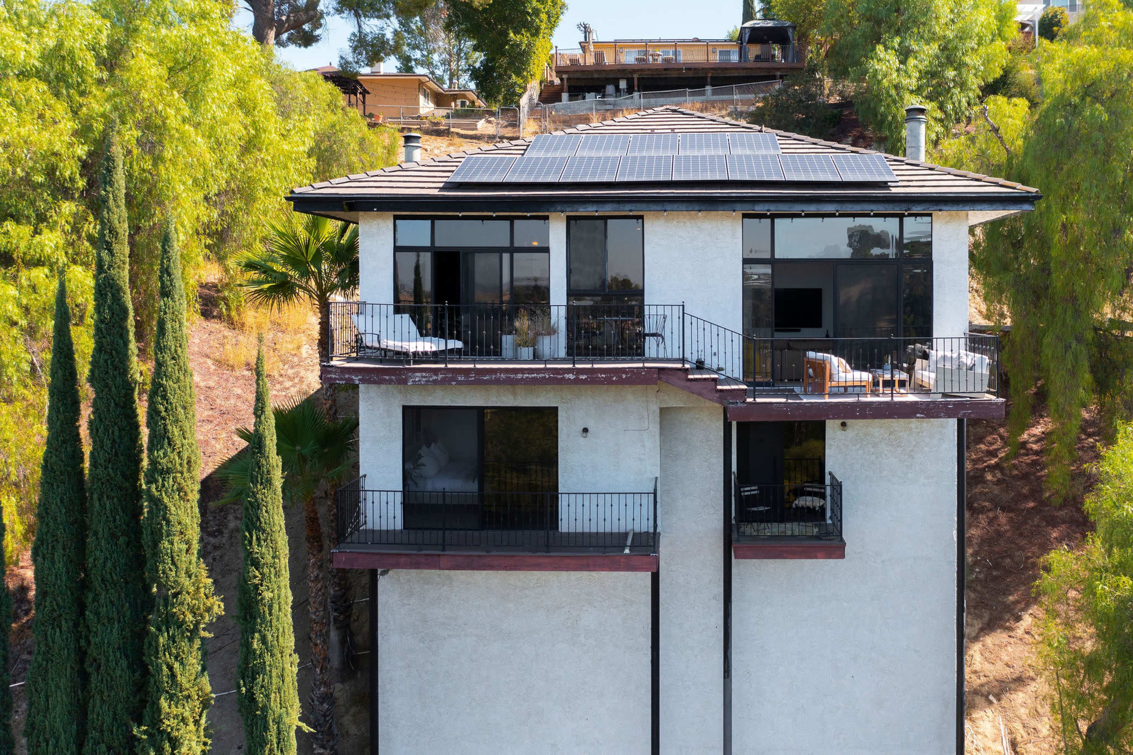 A modern two-story house with large windows, solar panels on the roof, and balconies overlooking a hillside.