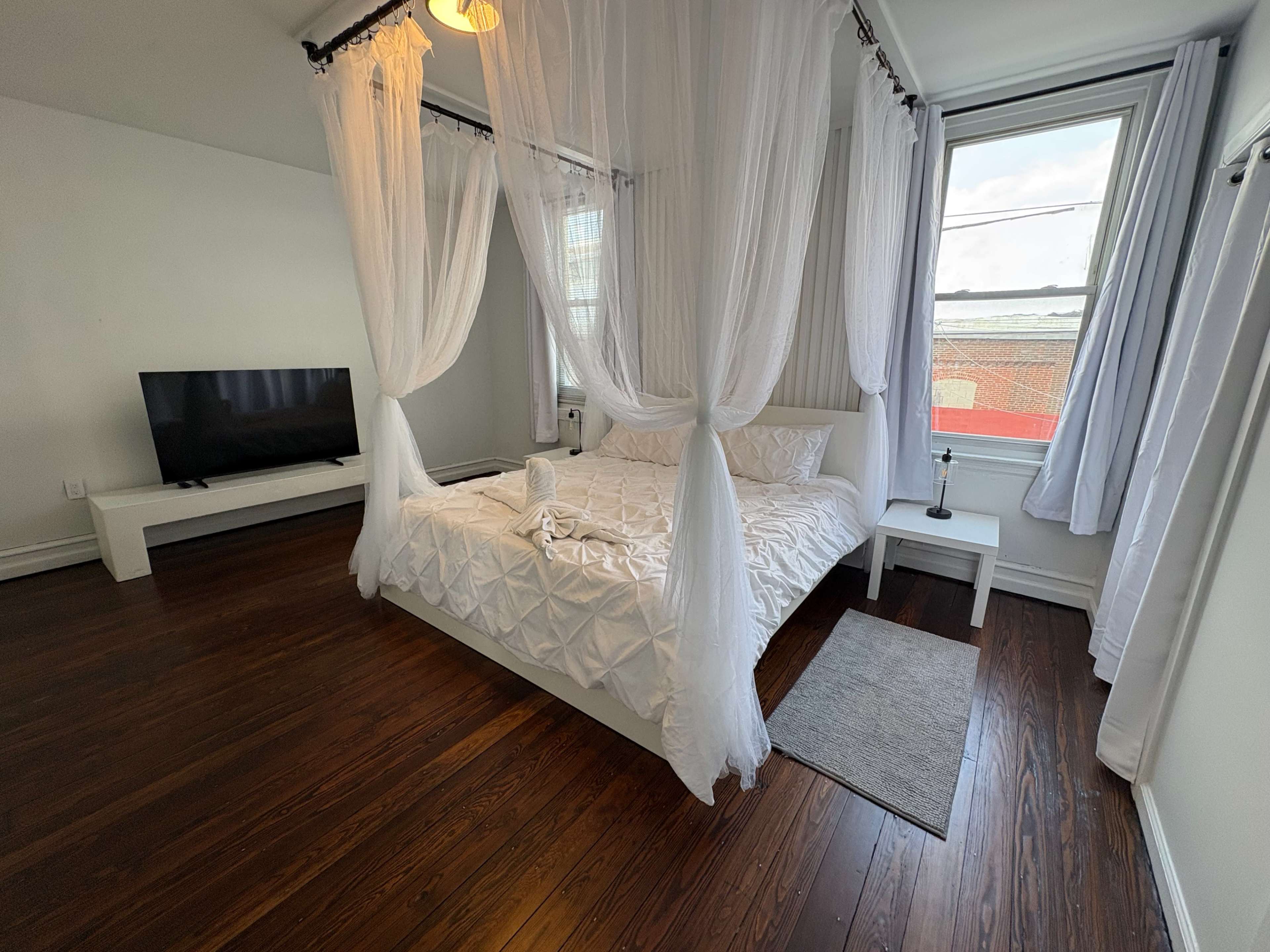 The image shows a modern bedroom featuring a four-poster bed with white drapes, a flat-screen television, and hardwood floors.