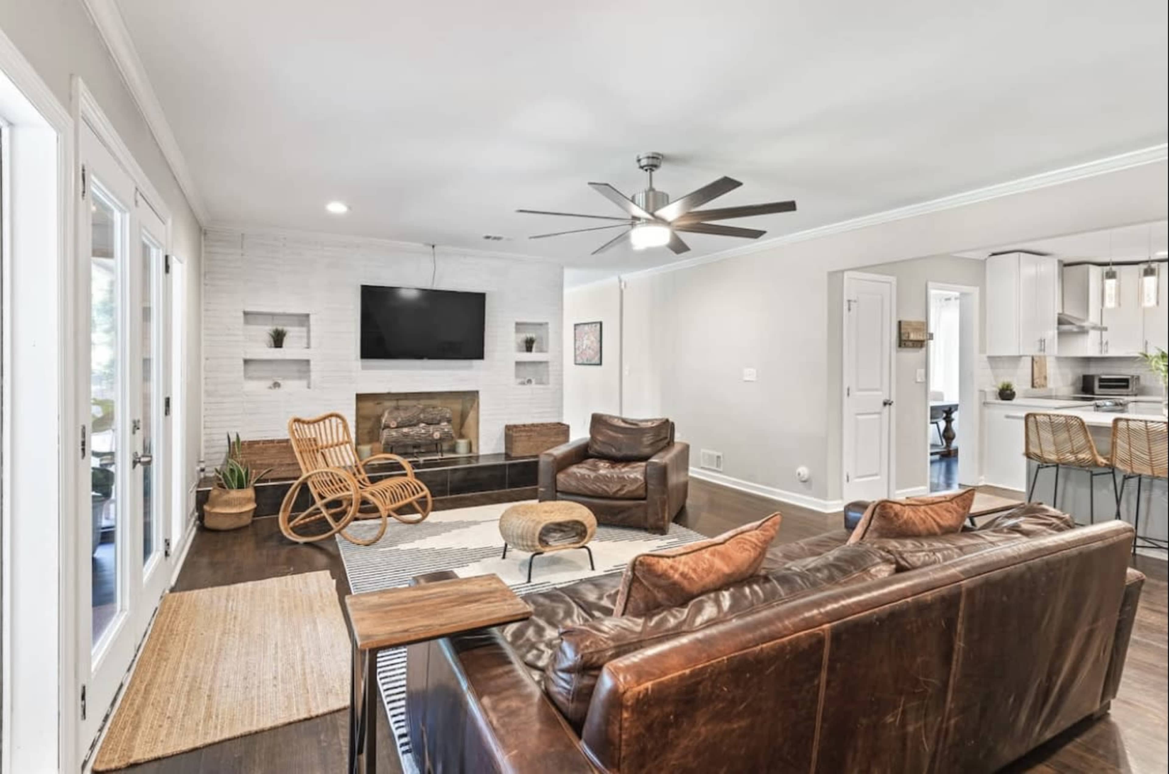 A modern living room with a brown leather sofa, a matching armchair, a wooden rocking chair, a wall-mounted television, and a light-colored brick fireplace.