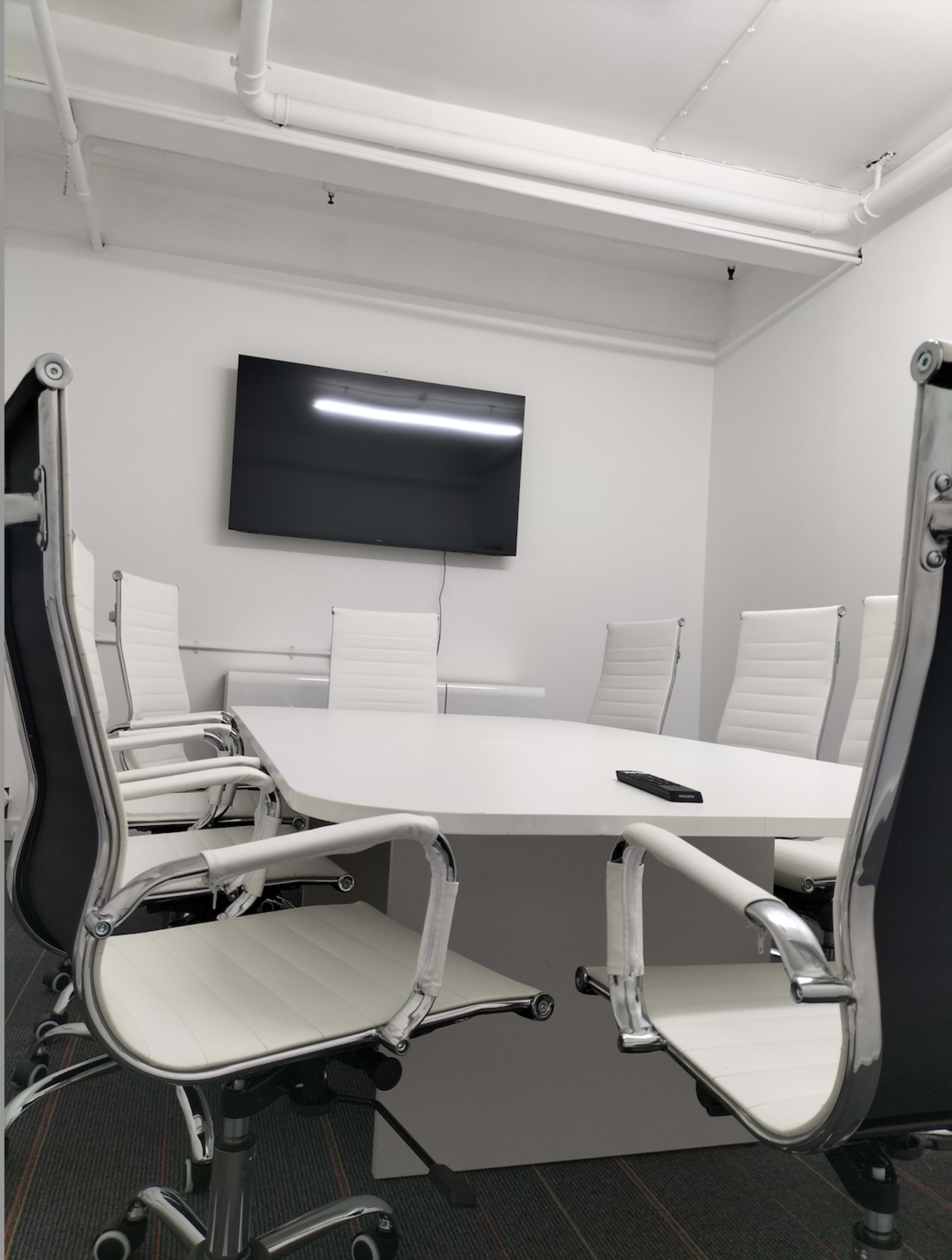 A modern conference room features a large white table surrounded by six white chairs and a wall-mounted television.
