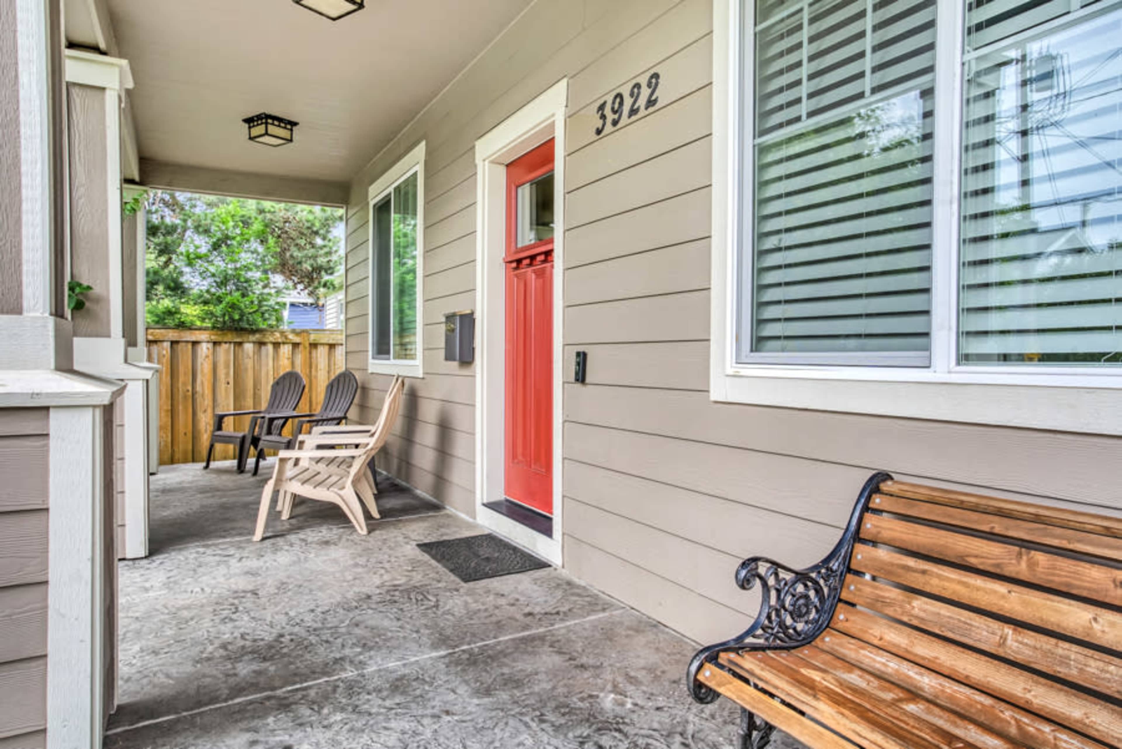 A covered porch with two wooden chairs and a bench, featuring a bright red front door and a fenced yard in the background.