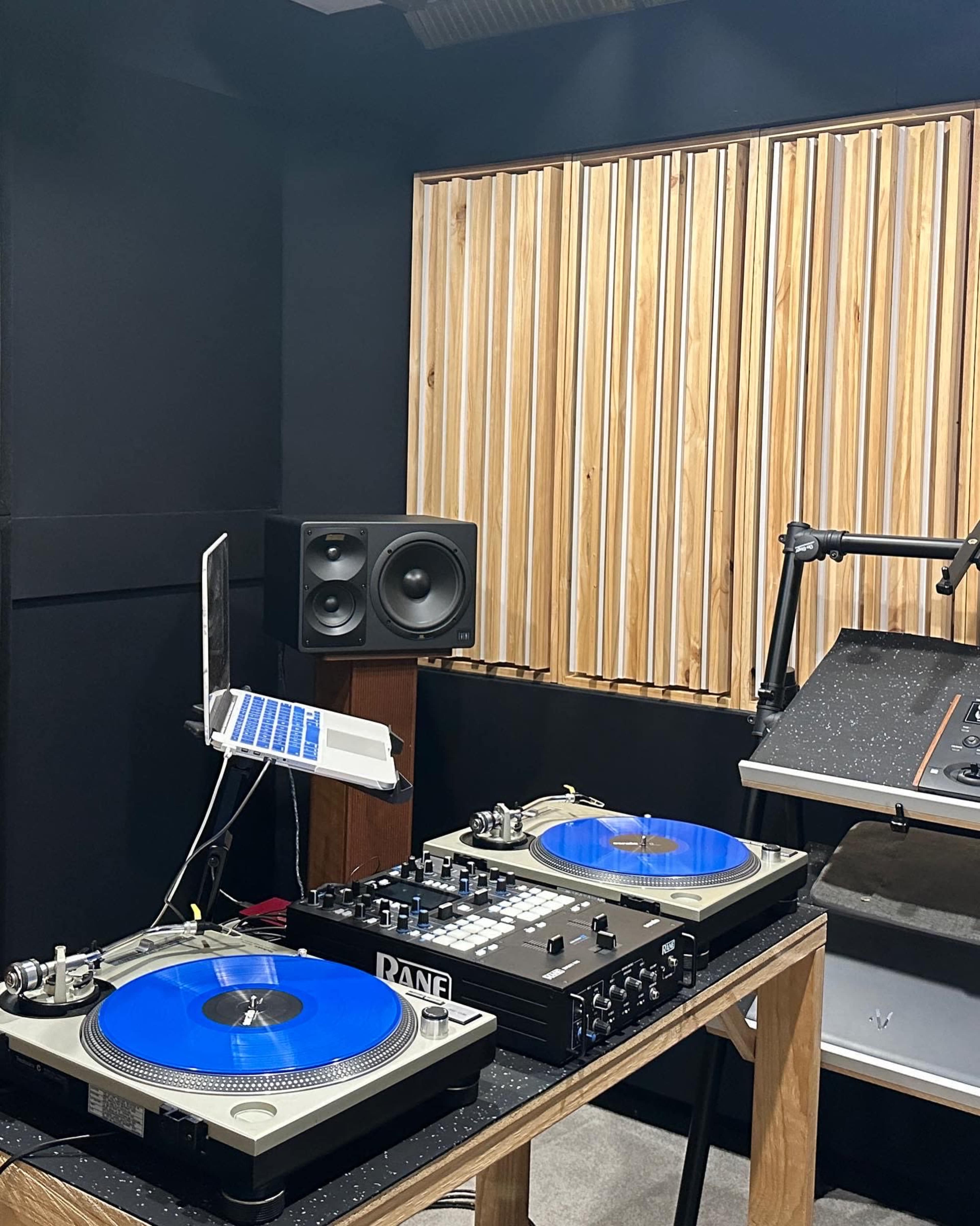 The image shows a DJ setup featuring two turntables with blue platters, a laptop, a mixer, and studio speakers in a soundproofed room.