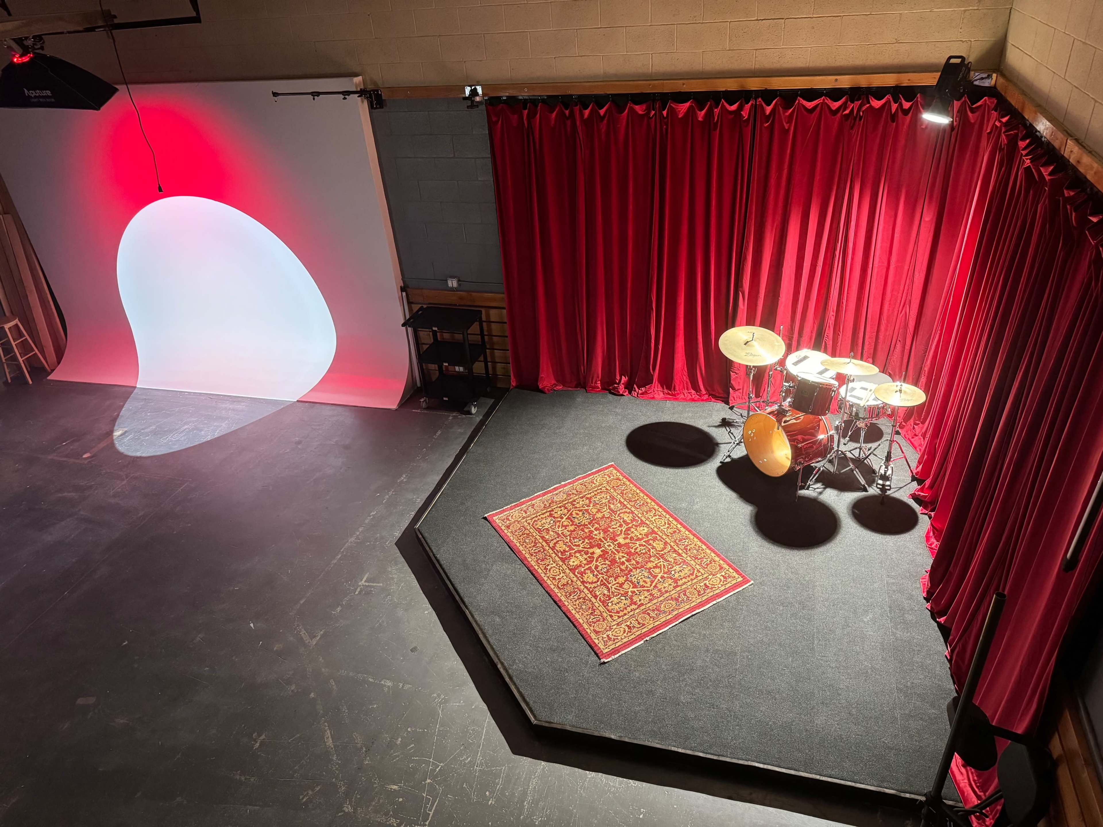The image shows a stage with a red curtain backdrop, a drum set on a black platform, and a decorative rug on the floor, illuminated by spotlights.