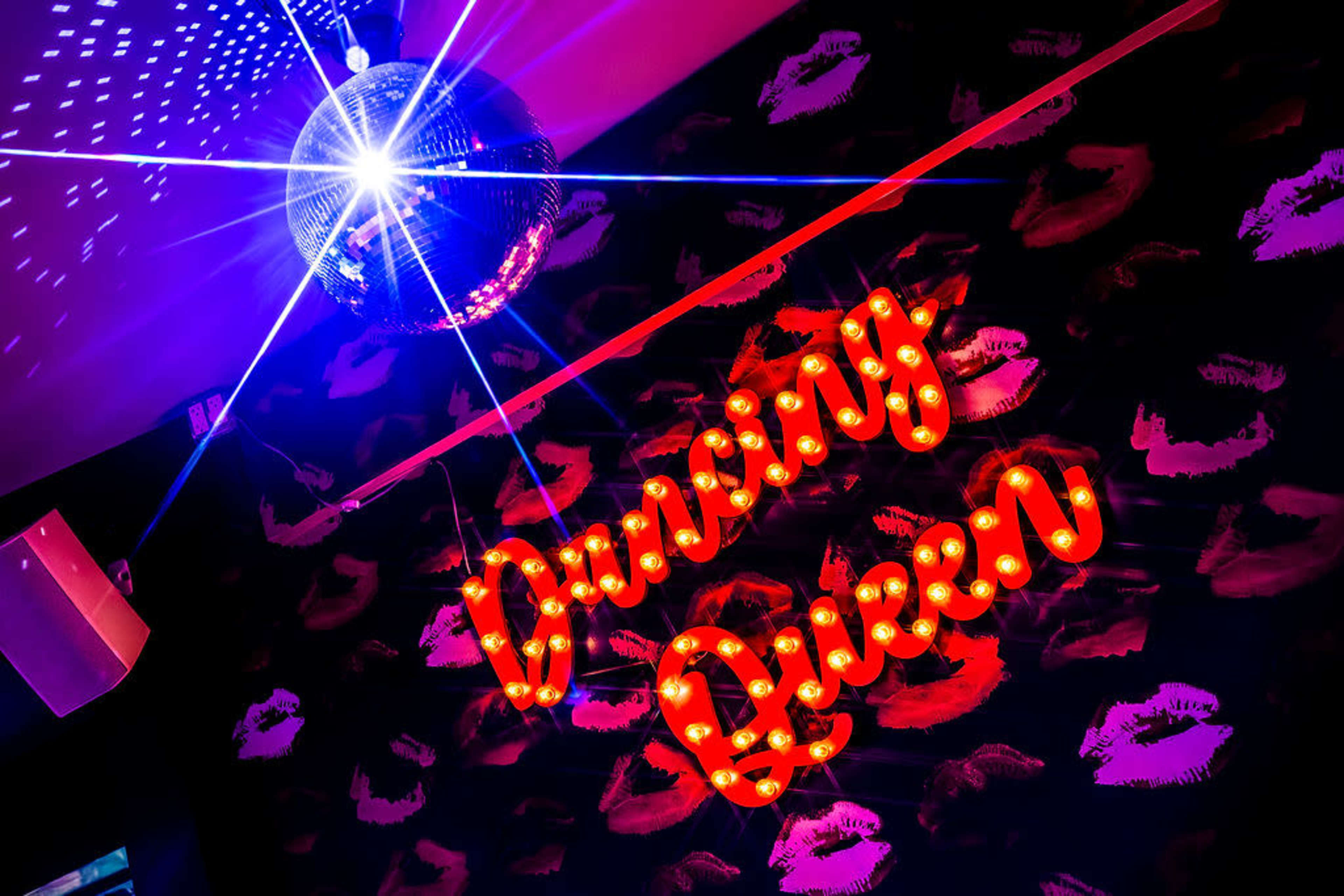 A mirrored disco ball hangs above the vibrant red "Dancing Queen" sign against a wall patterned with lips.
