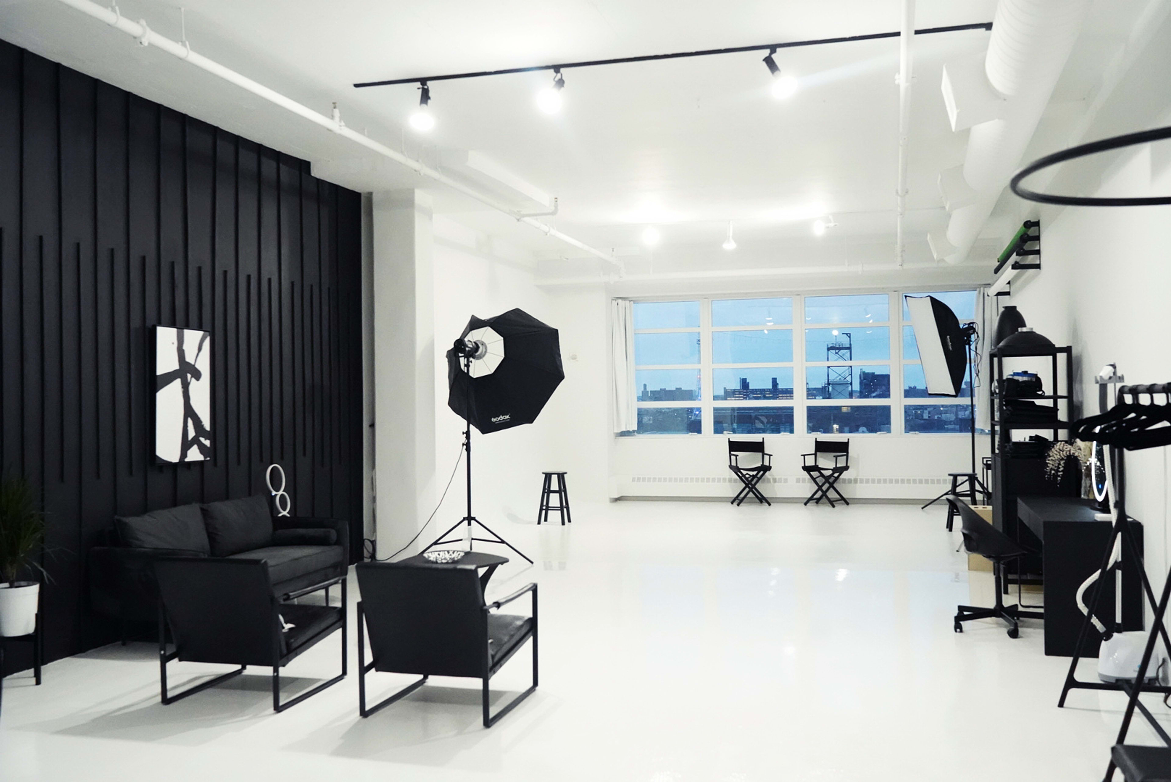A spacious, well-lit studio with black and white furniture, including chairs and a couch, and photography equipment set up near large windows.