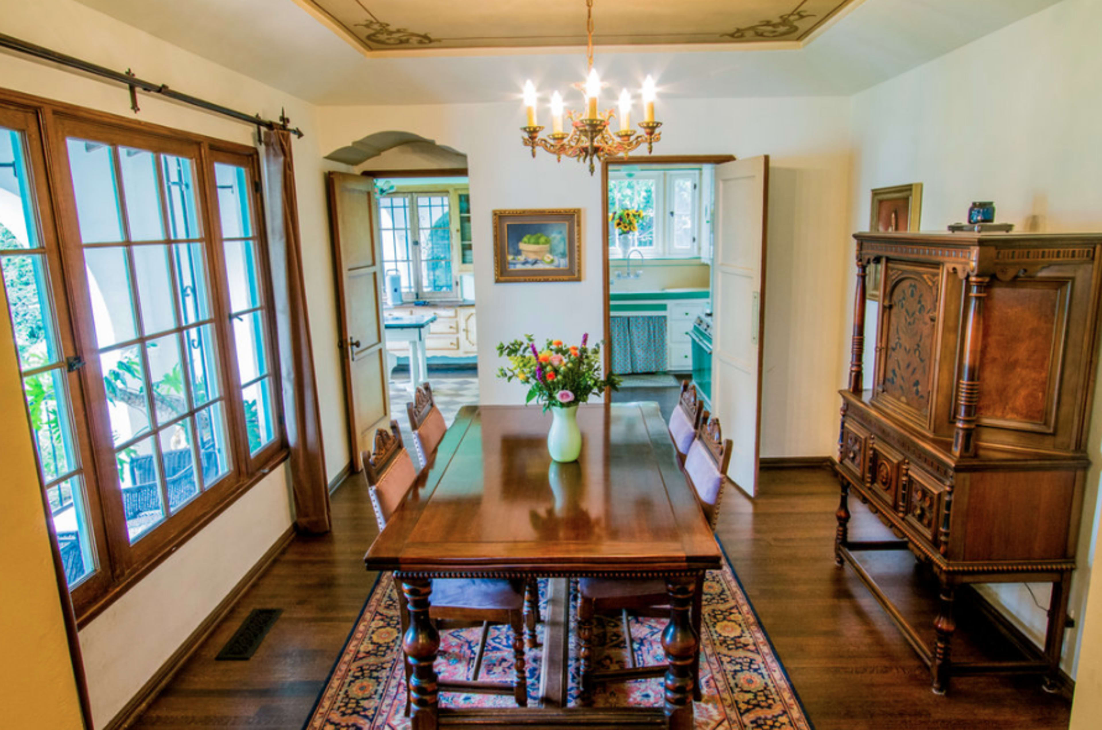 A dining room with a wooden table at the center, surrounded by chairs, and a chandelier above, with doorways leading to other areas of the home.