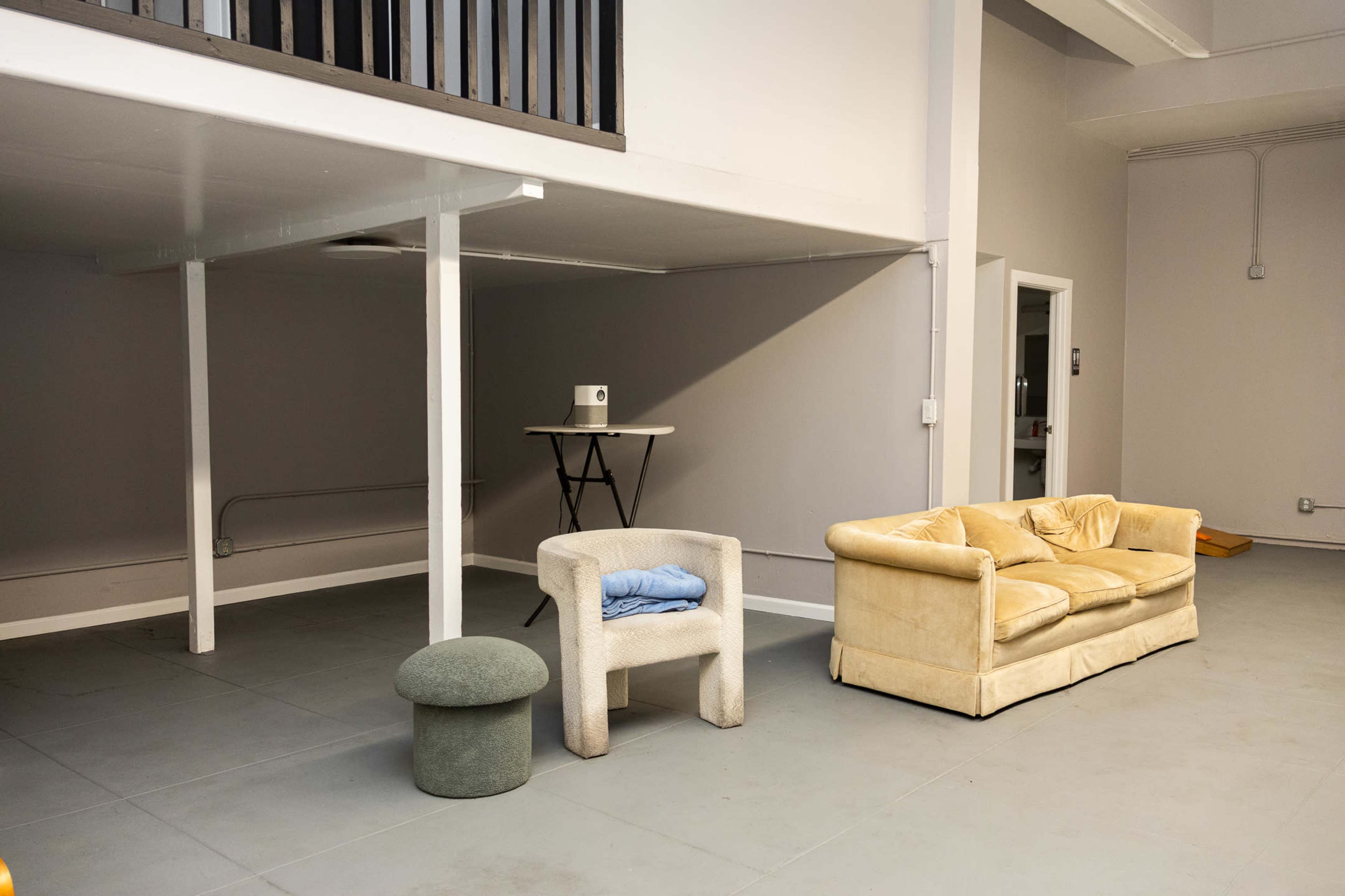 The image shows a minimalist indoor space featuring a yellow sofa, a white armchair, a green stool, and a folding table on a raised platform.