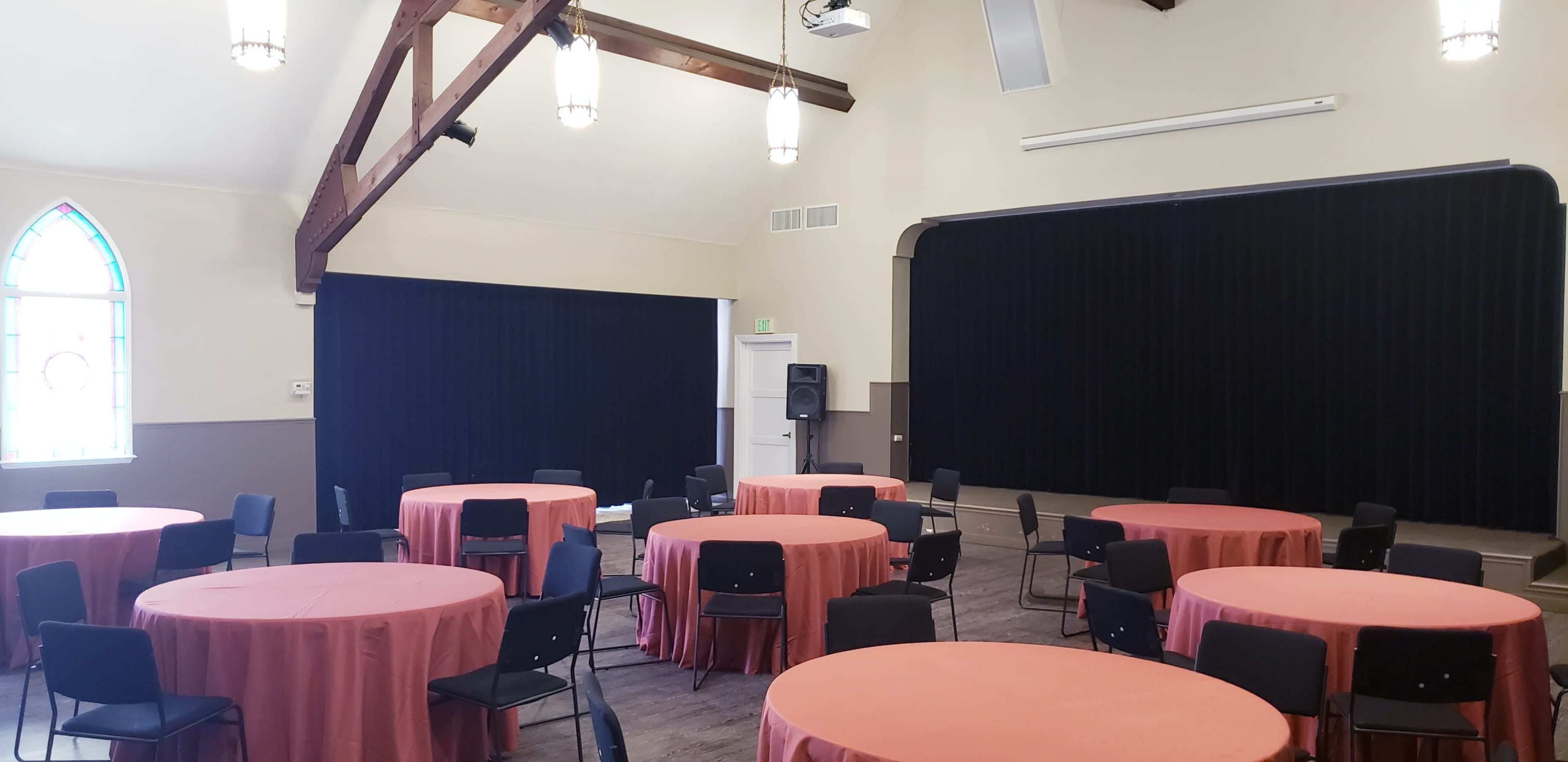 A spacious indoor hall with round tables covered in pink tablecloths, black chairs, and a dark stage at one end.