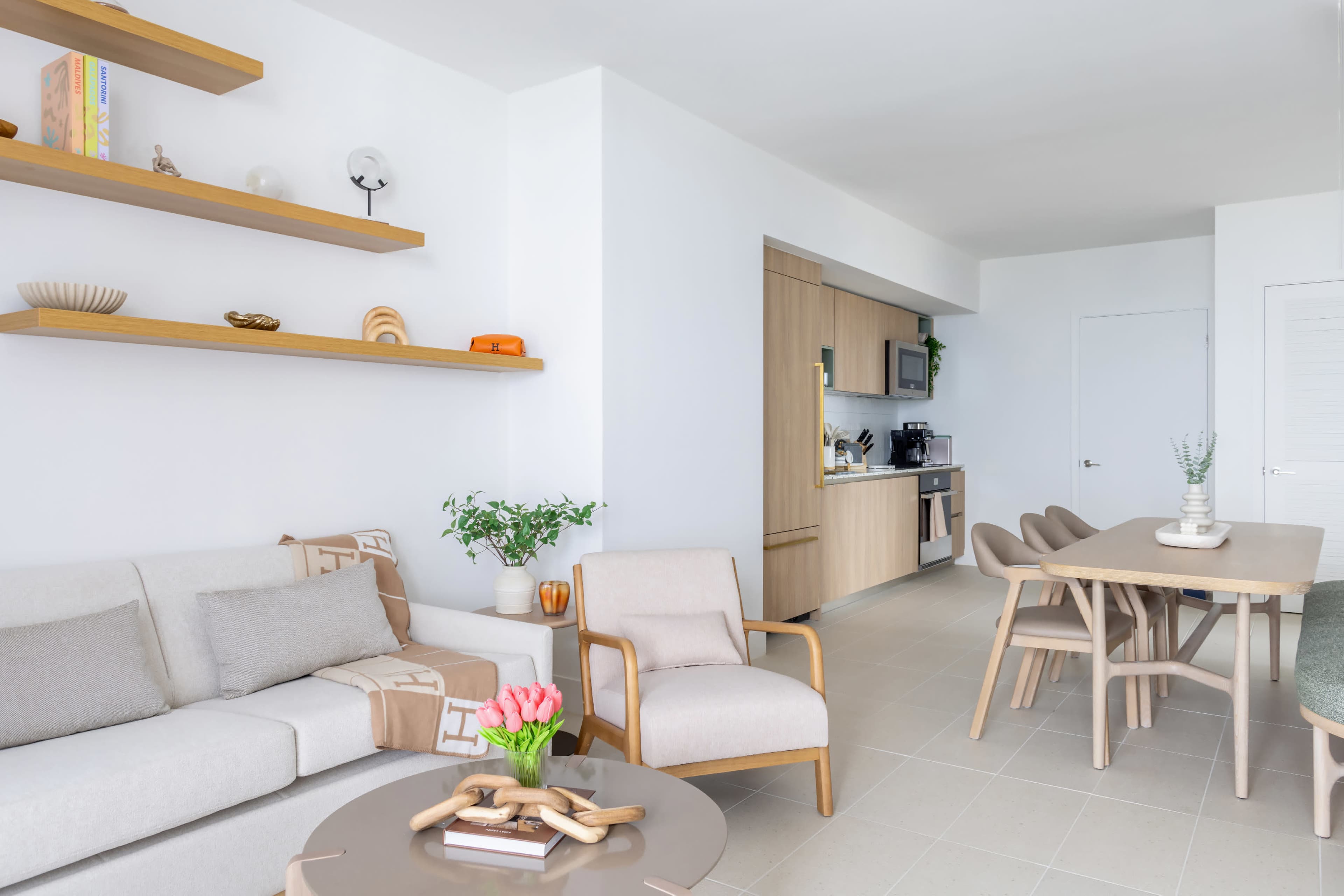 The image shows a modern living area featuring a beige sofa, a wooden dining table with chairs, and a kitchen area in the background with light cabinetry.