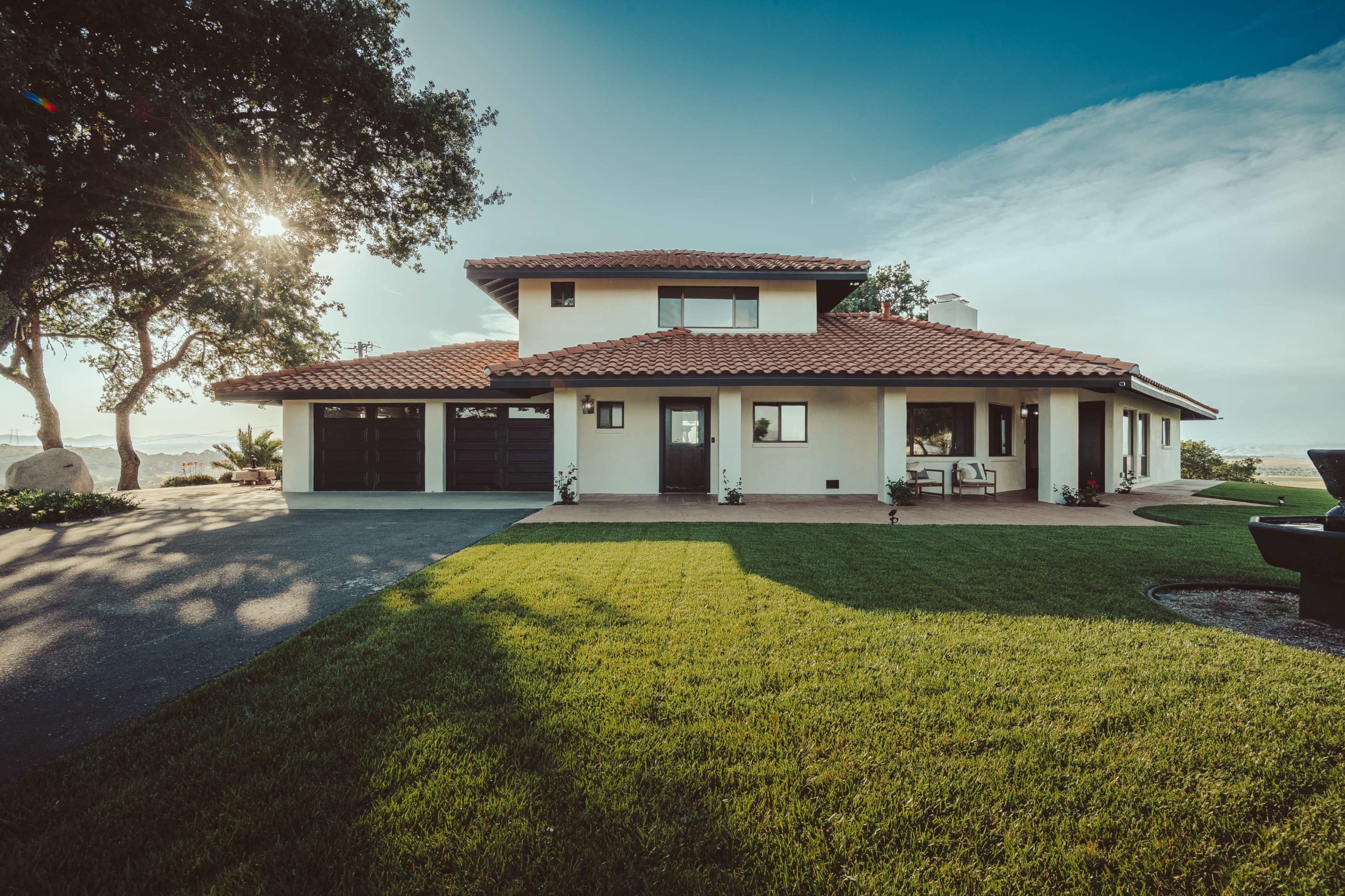 A two-story house with a red-tiled roof and a well-maintained lawn is situated under a clear blue sky.