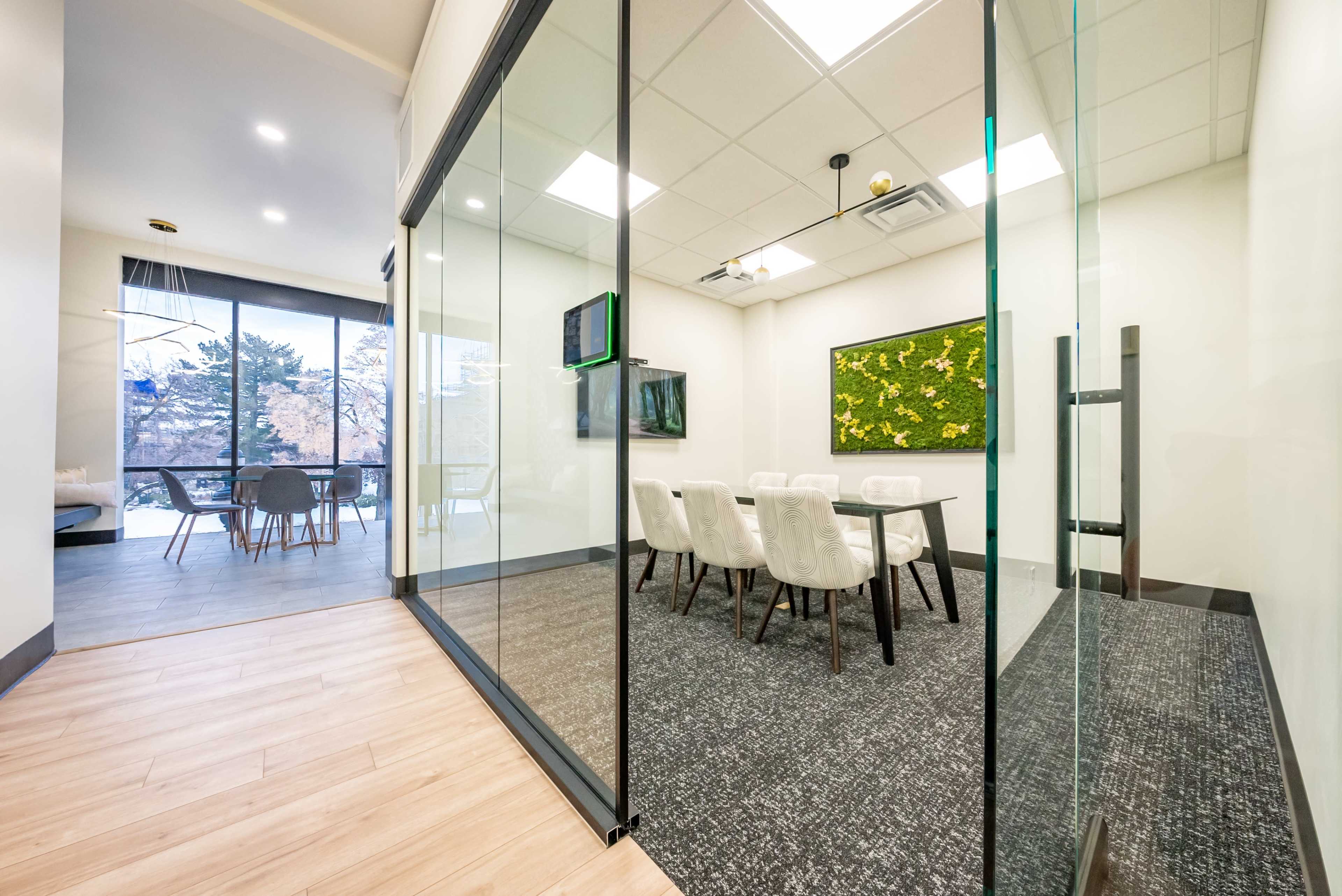 The image shows a modern conference room with glass walls, featuring a table and chairs, while a spacious area outside has large windows and additional seating.